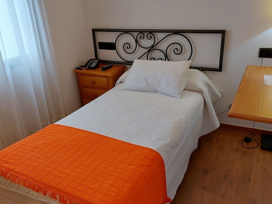 Una cama bien hecha con una manta blanca y naranja, una mesita de noche con un teléfono y un control remoto, y un aplique fijado a la pared.