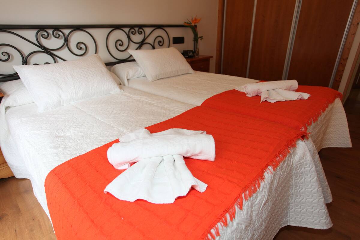 A double bed with white bedding and a red blanket, a nightstand with a vase of flowers, and a wooden floor.