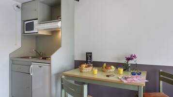 A compact kitchen with a microwave, sink, and a small refrigerator. There is a breakfast setup on a table with a basket of bread, a bowl of fruit, and a glass of orange juice.