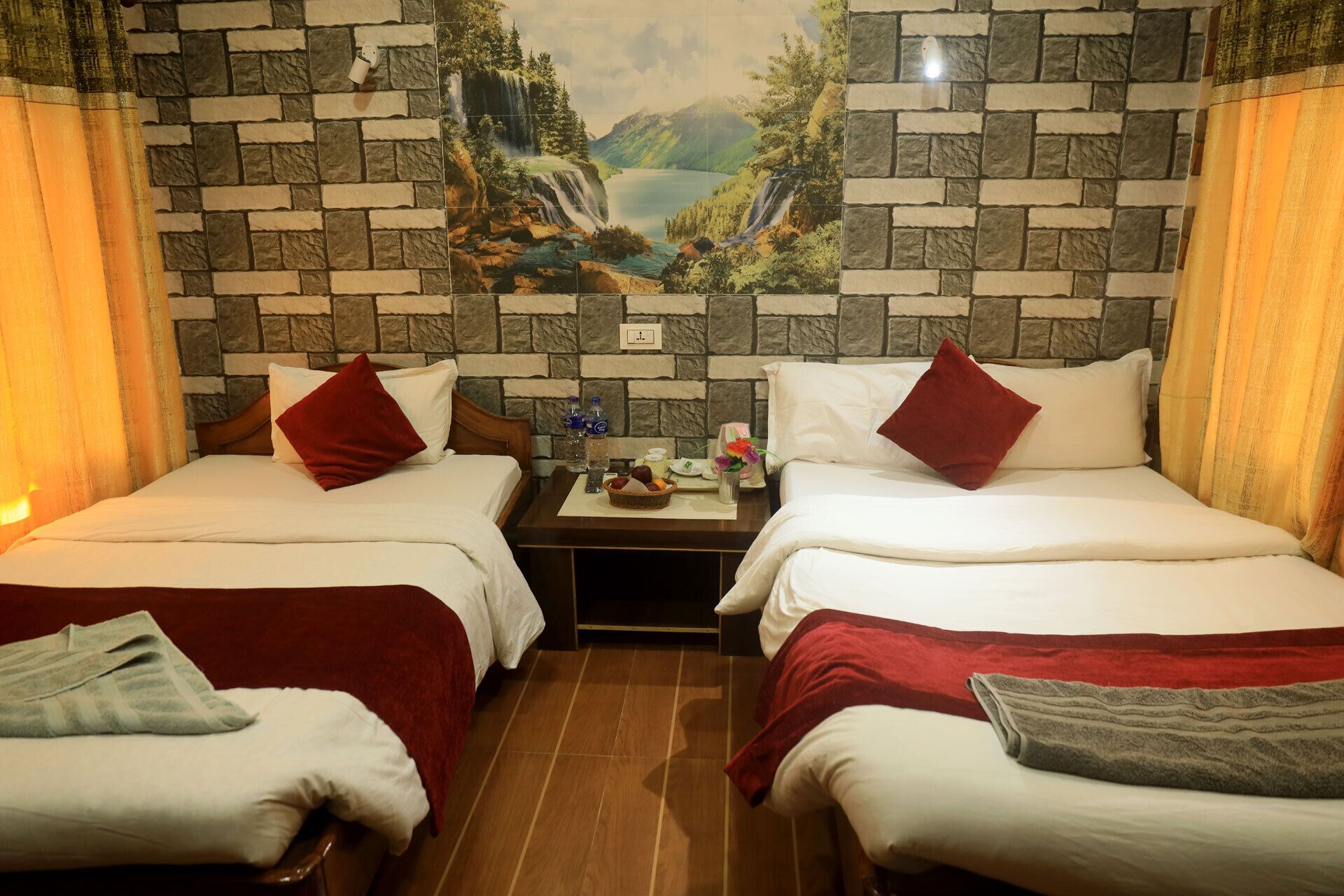 A hotel room with two beds, each with red pillows and white linens. A small table between the beds holds a basket of fruit and a bottle of water. The room features a stone wall with a scenic landscape mural.