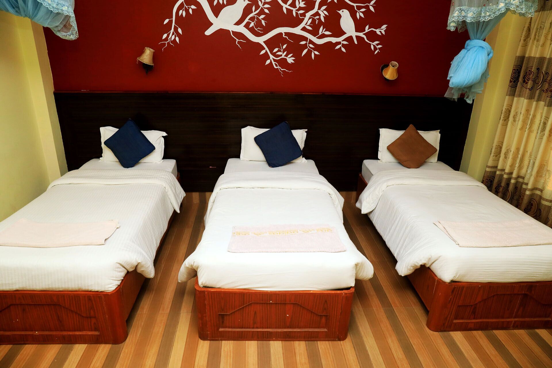 Three single beds with wooden frames and white bedding, a red wall with a white tree design, and a wooden floor.