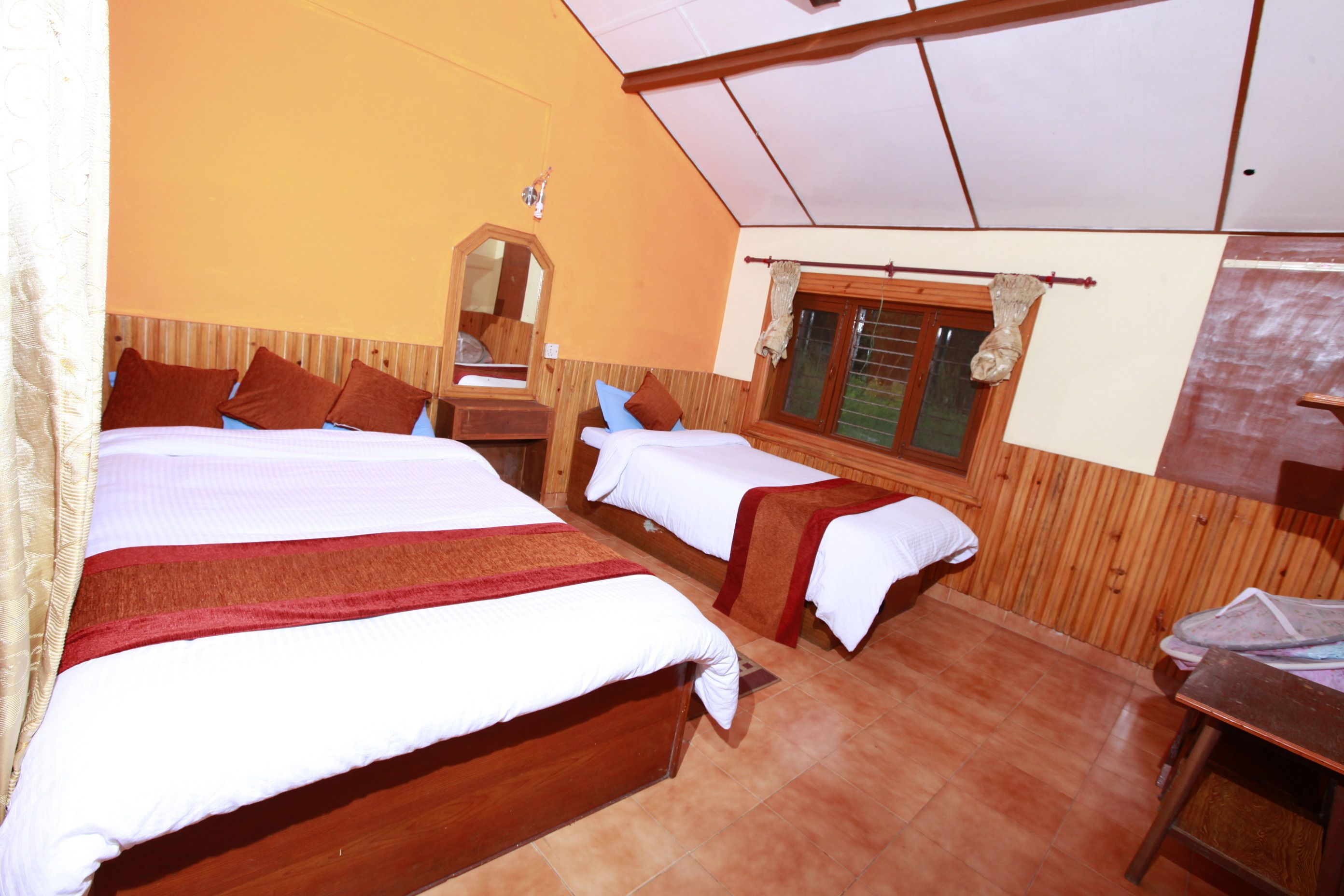 Ein Zimmer mit zwei Betten, Holz-Kopfteilen, einem Holztisch, einem Fenster mit Vorhängen und einem Spiegel an der Wand.