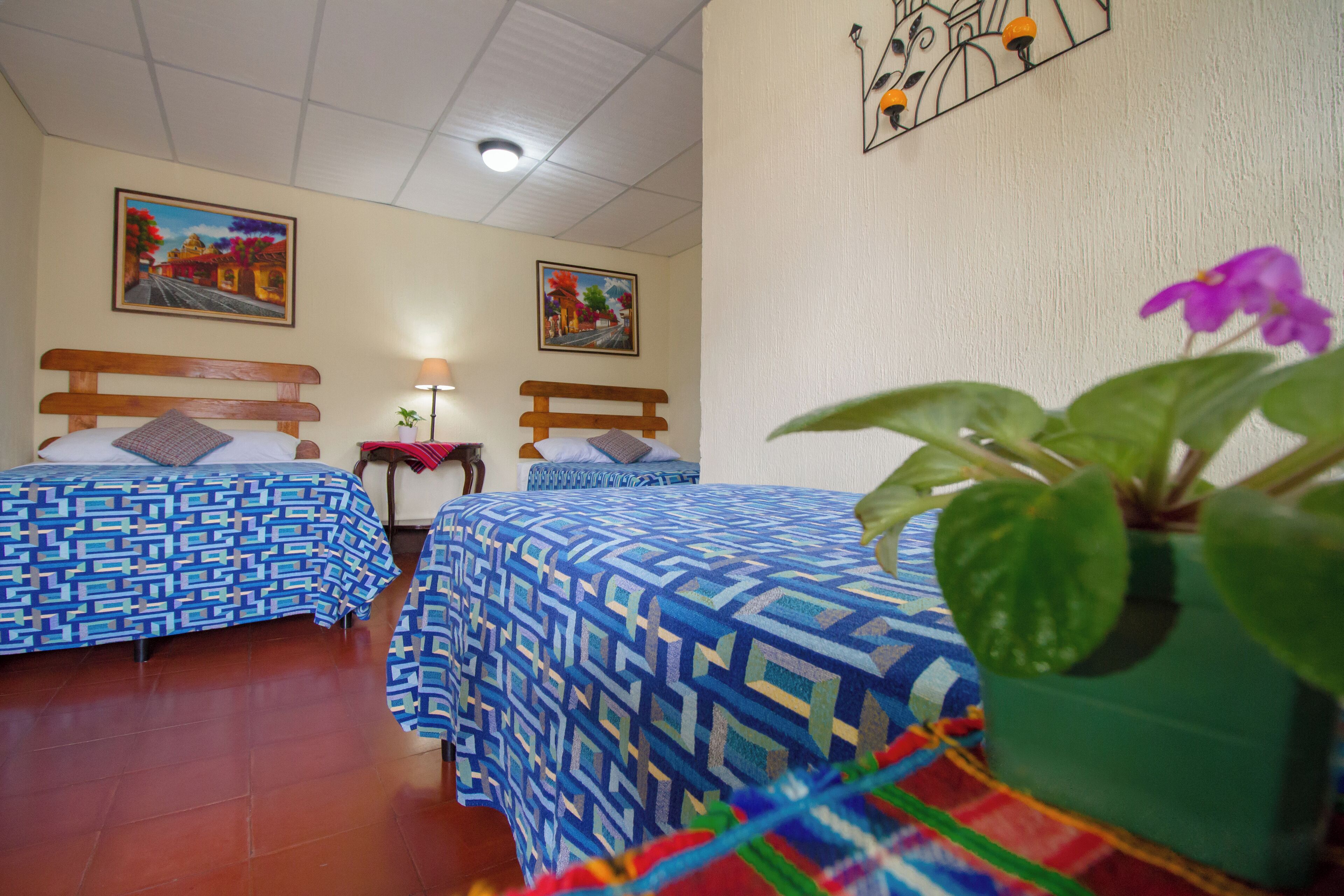 A hotel room with two beds, a nightstand, a lamp, and a wall with two framed pictures.