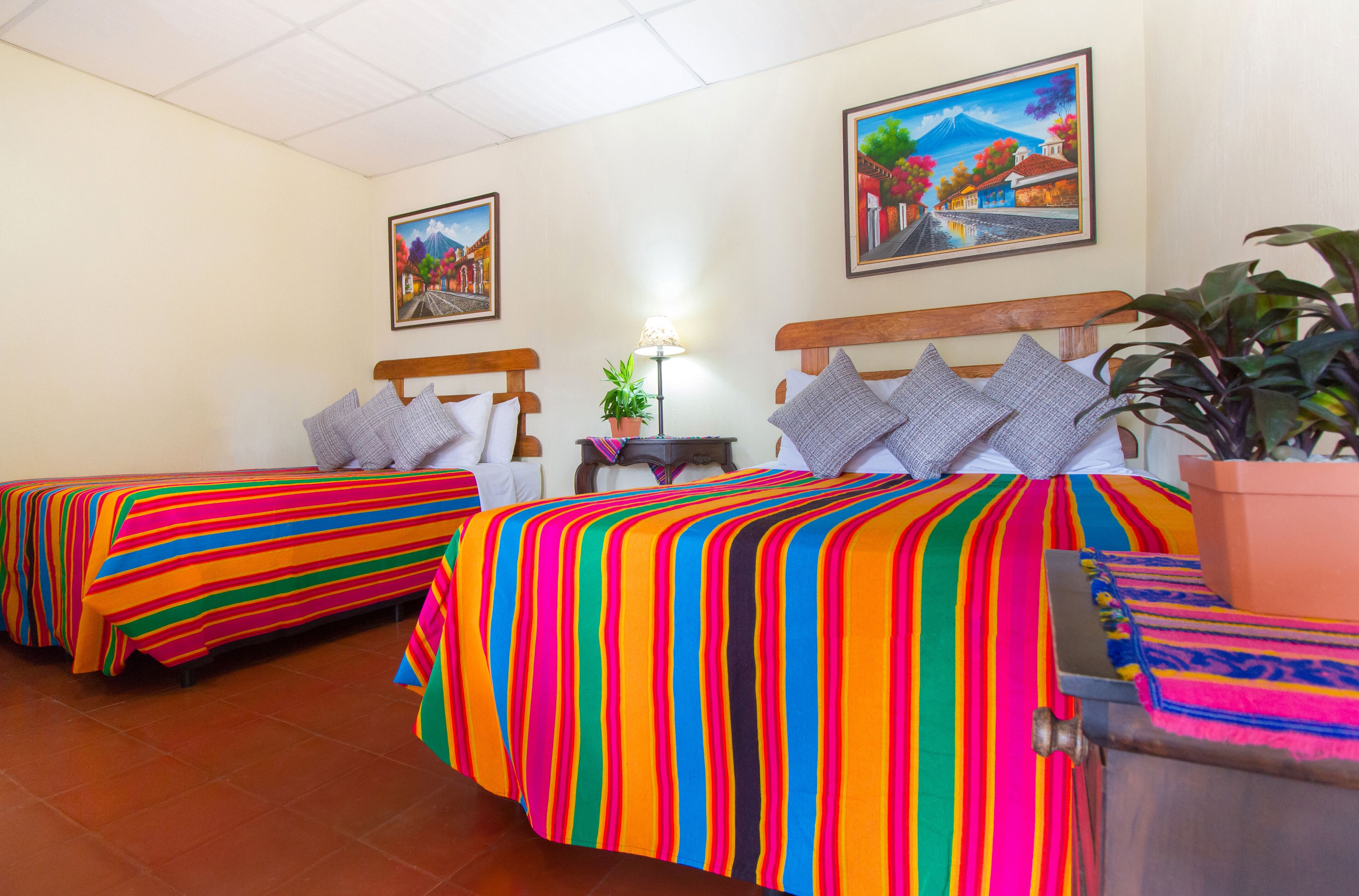 Une chambre d’hôtel avec deux lits, chacun agrémenté d’une literie à rayures colorées, une petite table avec une lampe et deux tableaux accrochés aux murs.