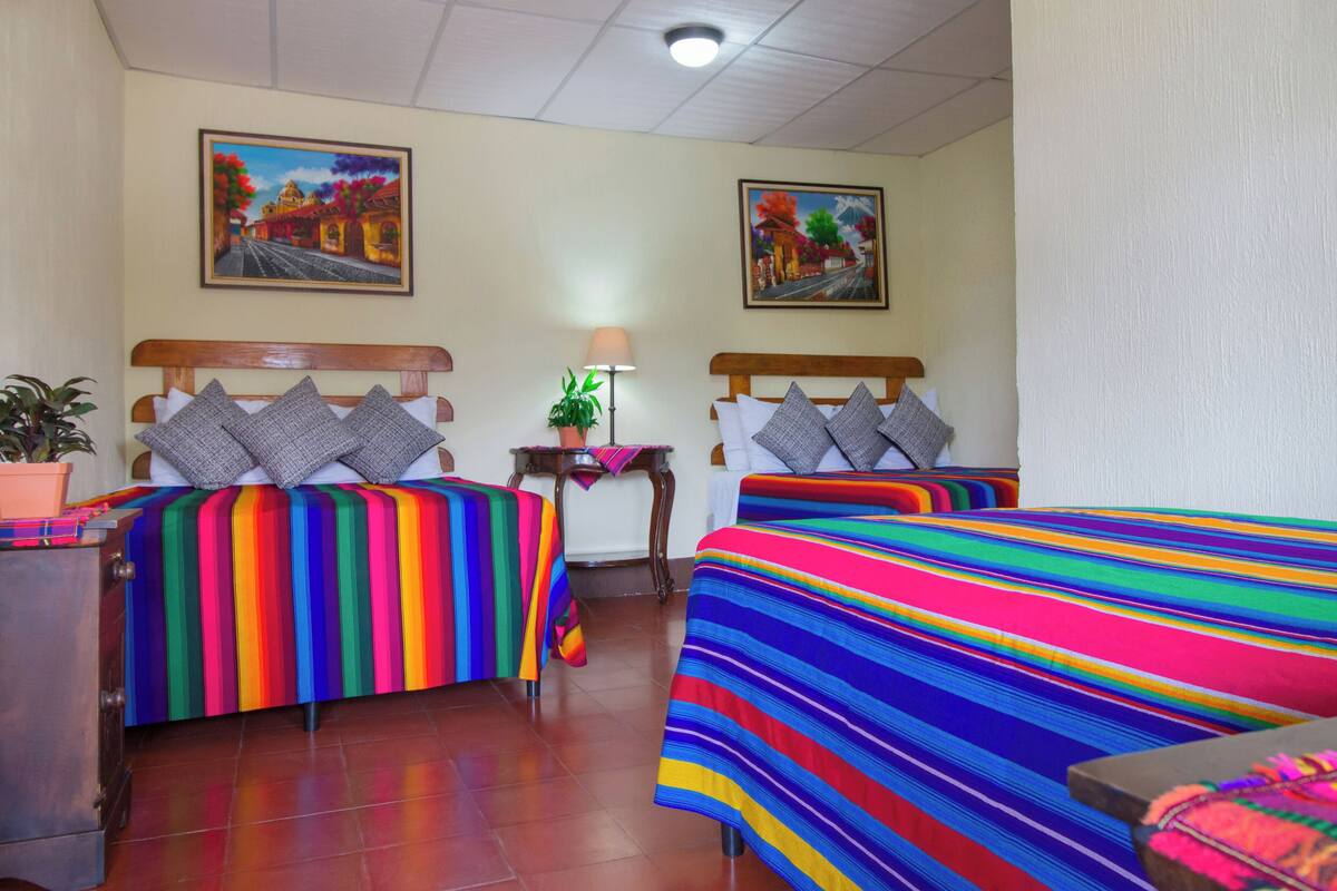 A hotel room with two beds, each with a colorful striped bedspread. There are two paintings on the walls, a small table with a lamp, and a potted plant.