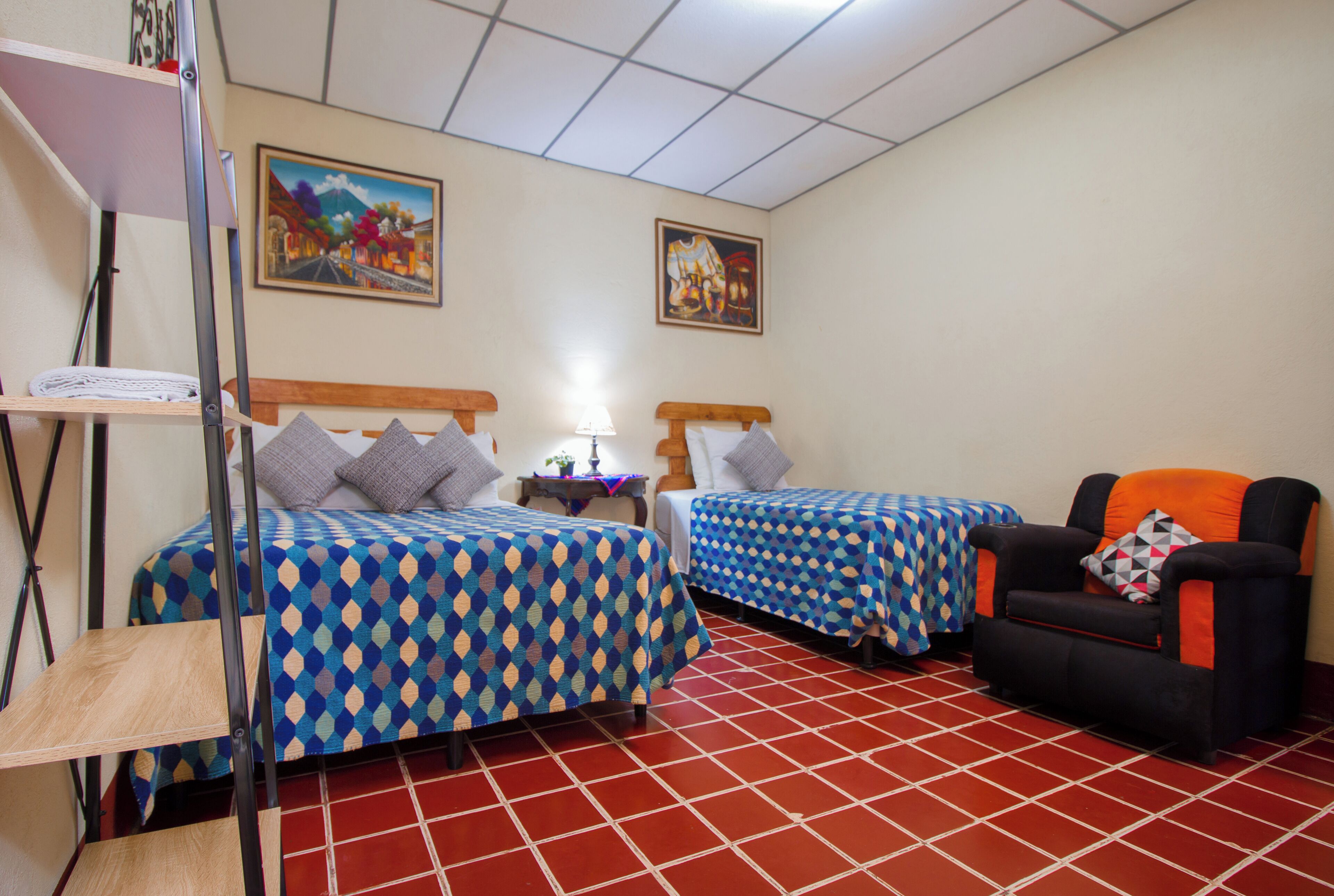 A hotel room with two beds, a chair, a shelf, and two paintings on the wall.