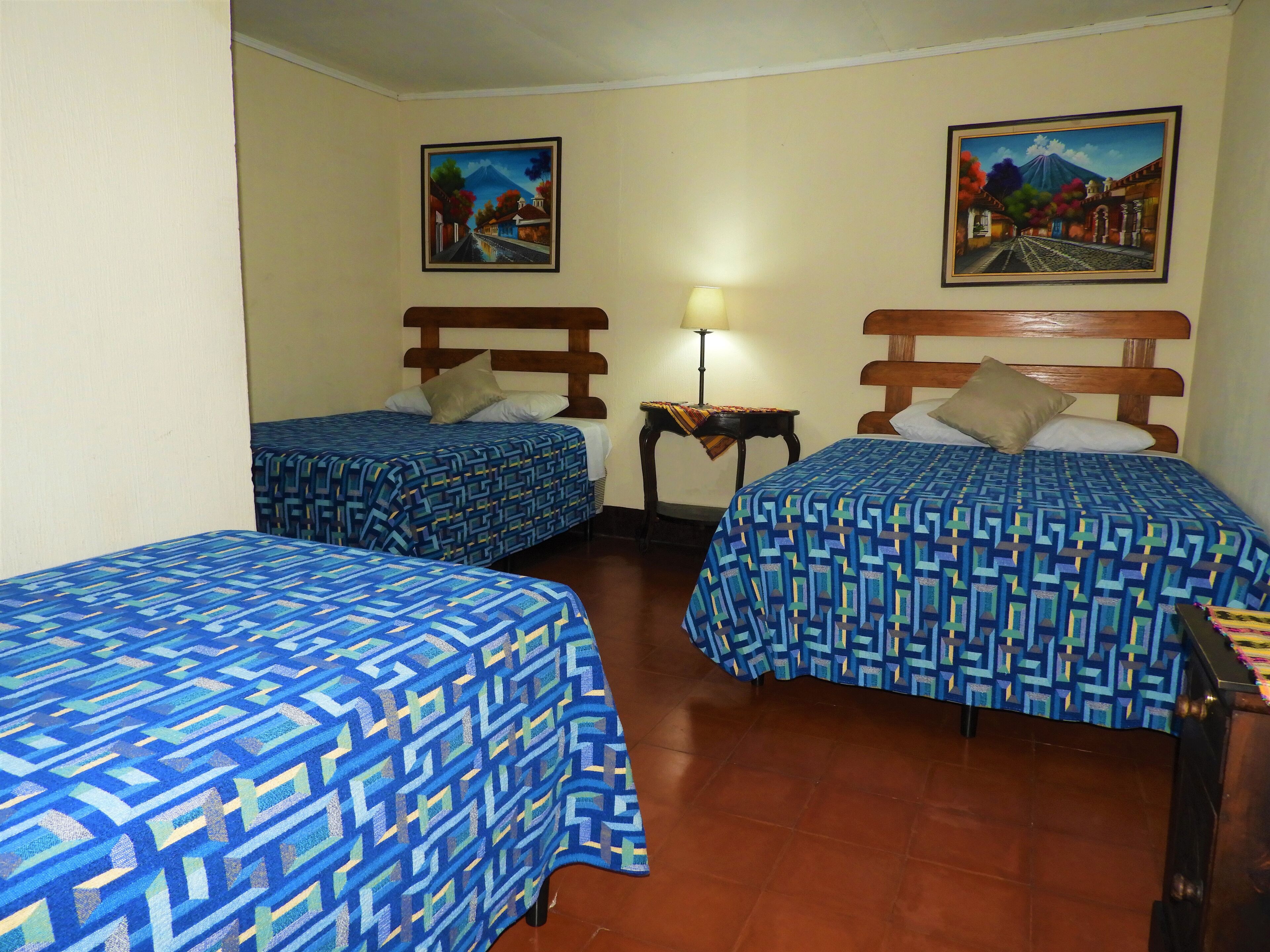 A hotel room with two beds, each with a patterned bedspread, a nightstand with a lamp, and two paintings on the walls.