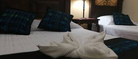 Habitación de hotel con dos camas, cada una con una colcha blanca y almohadas a cuadros azules. Una toalla blanca doblada está colocada en el suelo entre las camas.