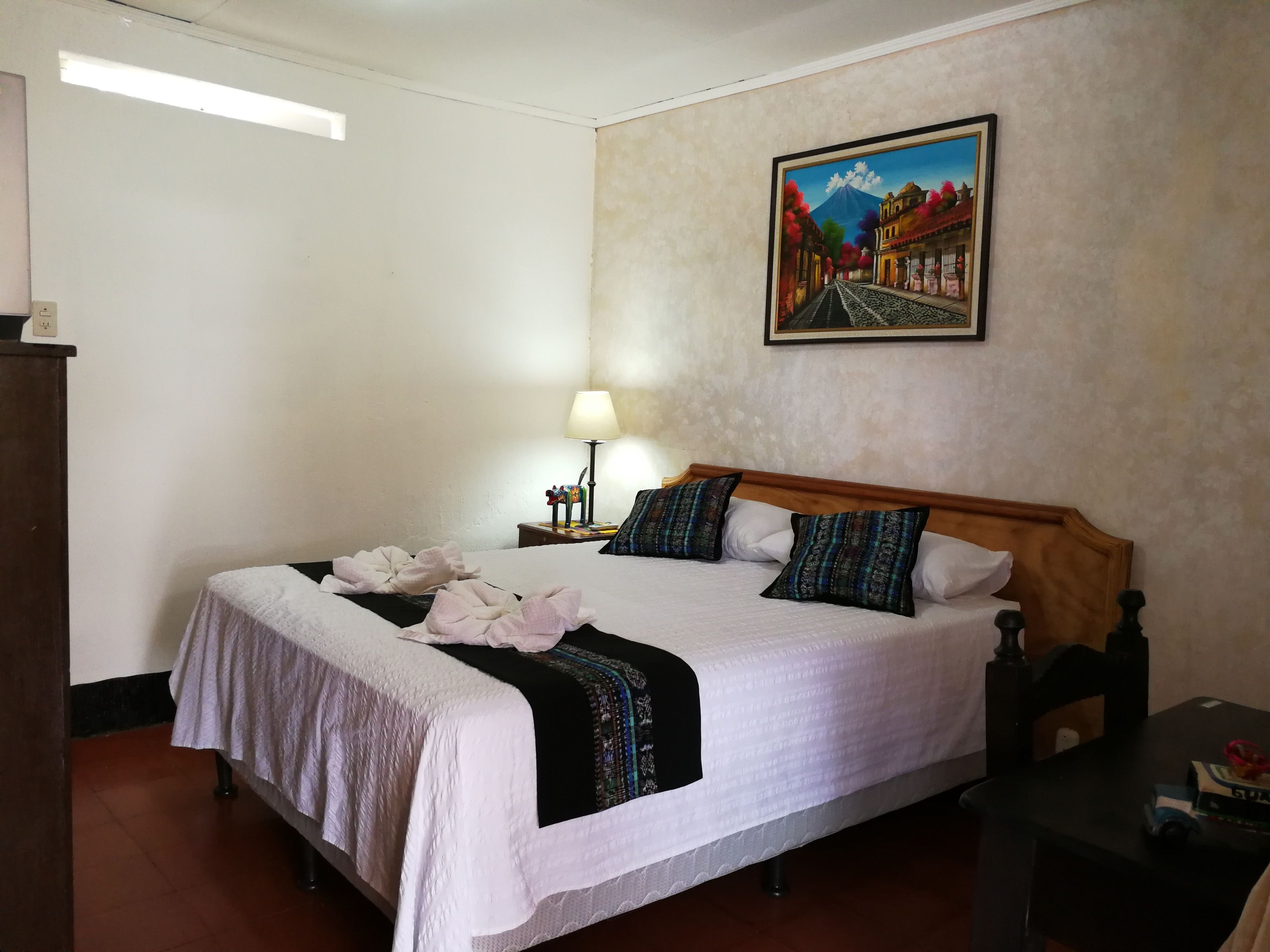 A neatly made bed with a white comforter and two patterned pillows, a bedside lamp, and a framed painting on the wall.
