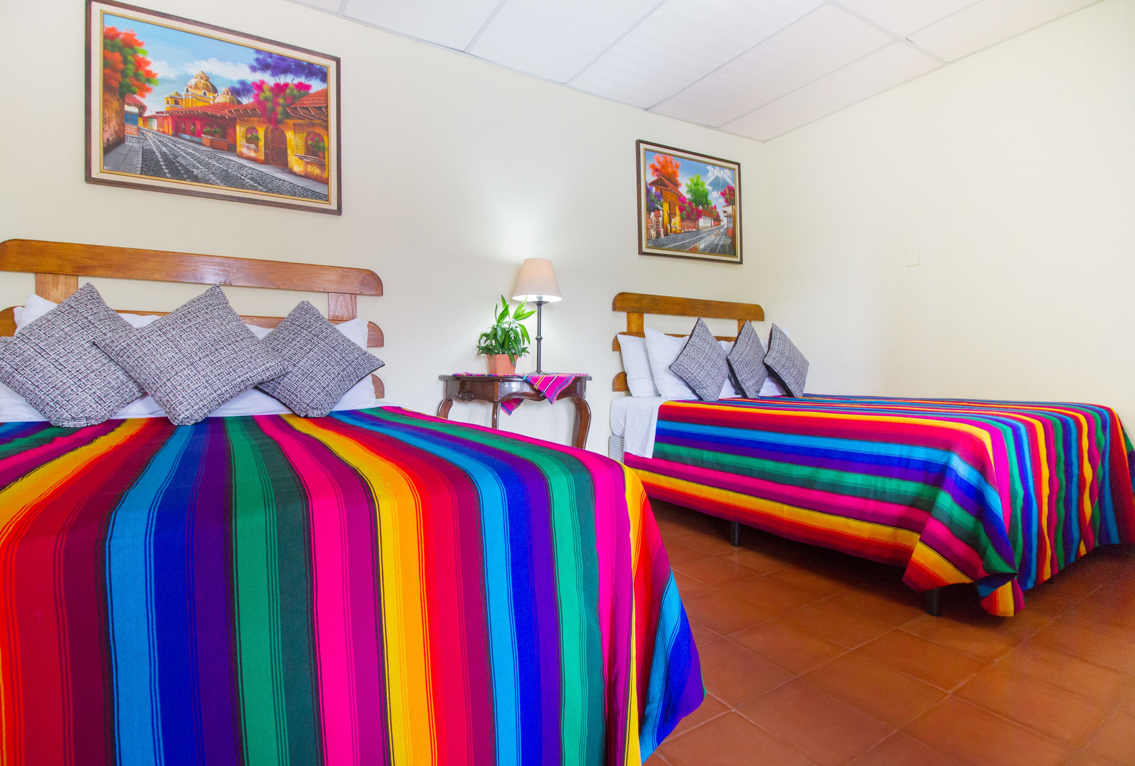 Habitación de hotel con dos camas, cada una con una colcha a rayas y colores vivos. Hay dos cuadros en las paredes, una lámpara sobre una mesita de noche y una pequeña planta en maceta.