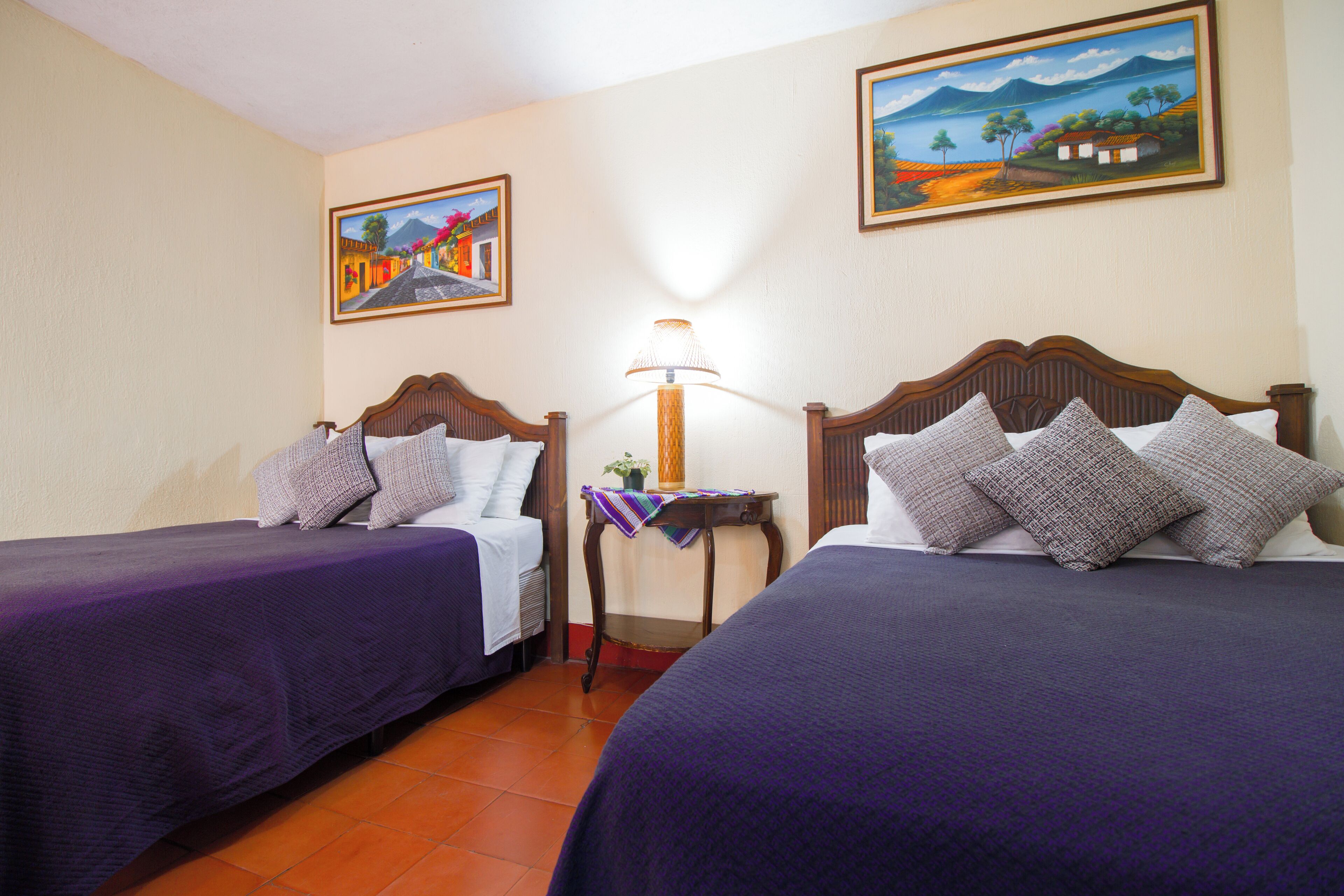 Habitación de hotel con dos camas, una mesita de noche y una lámpara. Hay pinturas en las paredes.