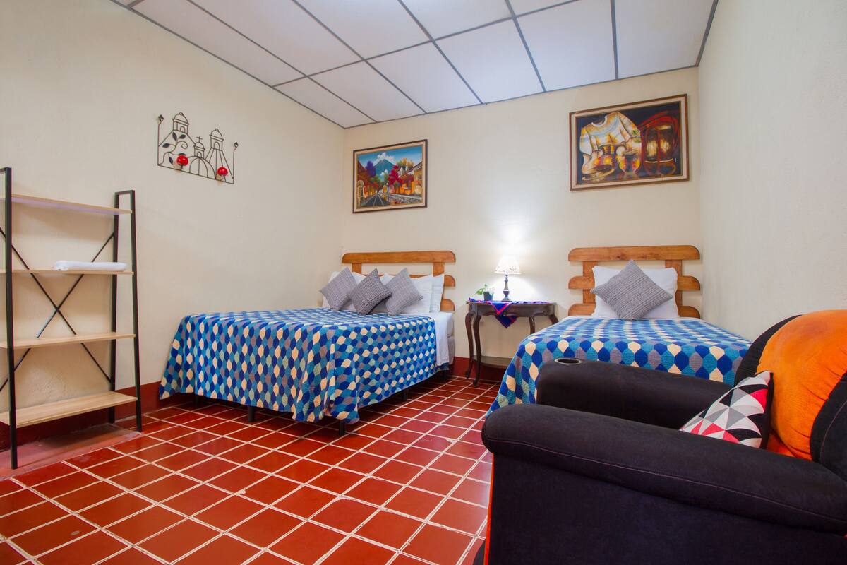 A room with two beds, a chair, a shelf, and two paintings on the walls.