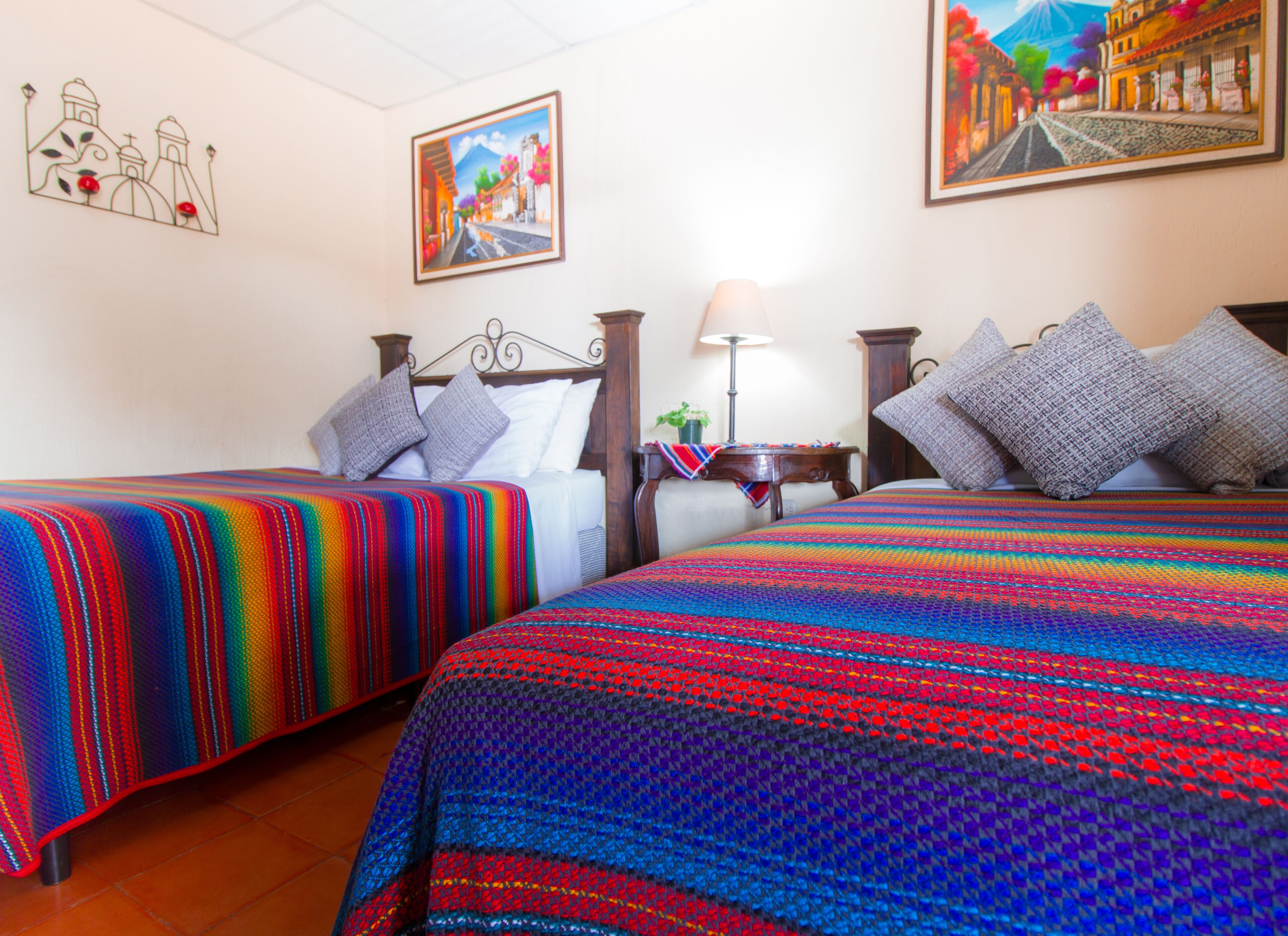 Ein Hotelzimmer mit zwei Betten, jeweils mit farbig gestreiften Bettdecken. Ein Nachttisch mit Lampe und ein kleiner Tisch mit einer Pflanze sind vorhanden. Das Zimmer hat ein schlichtes, aufgeräumtes Design mit weißen Wänden und einer Deckenleuchte.