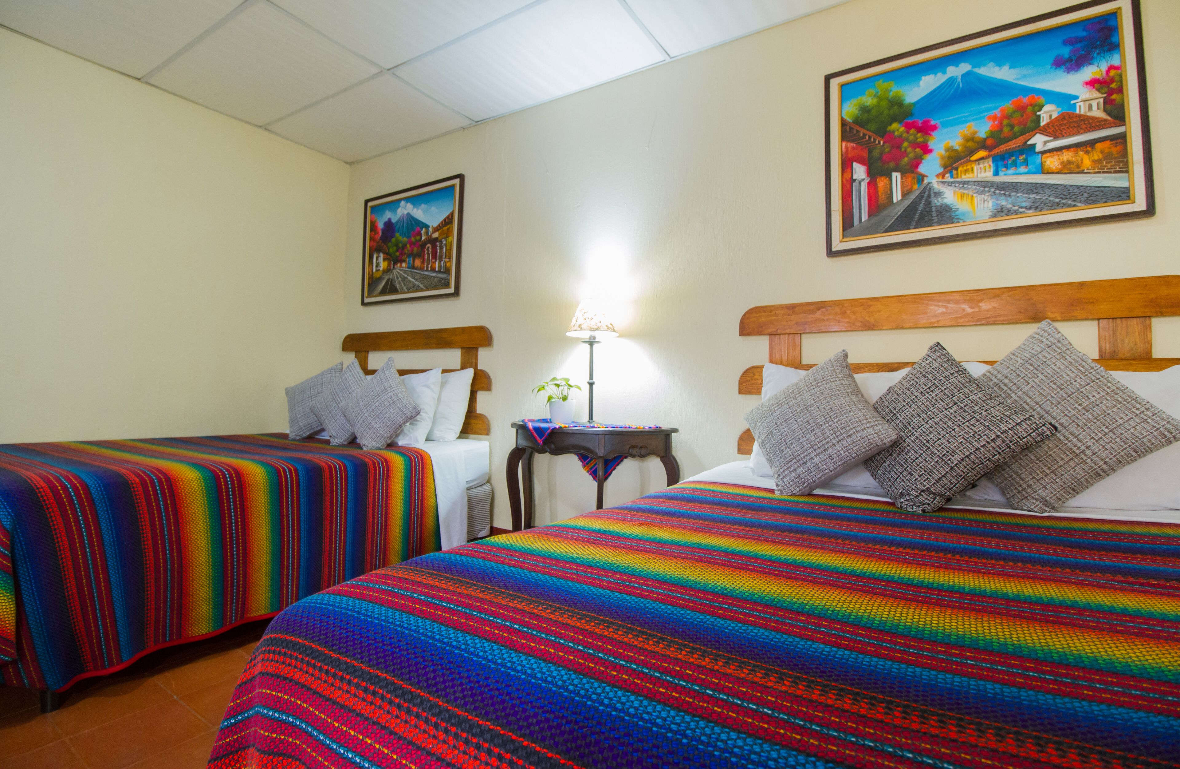 A hotel room with two beds, a nightstand, a lamp, and two paintings on the walls.