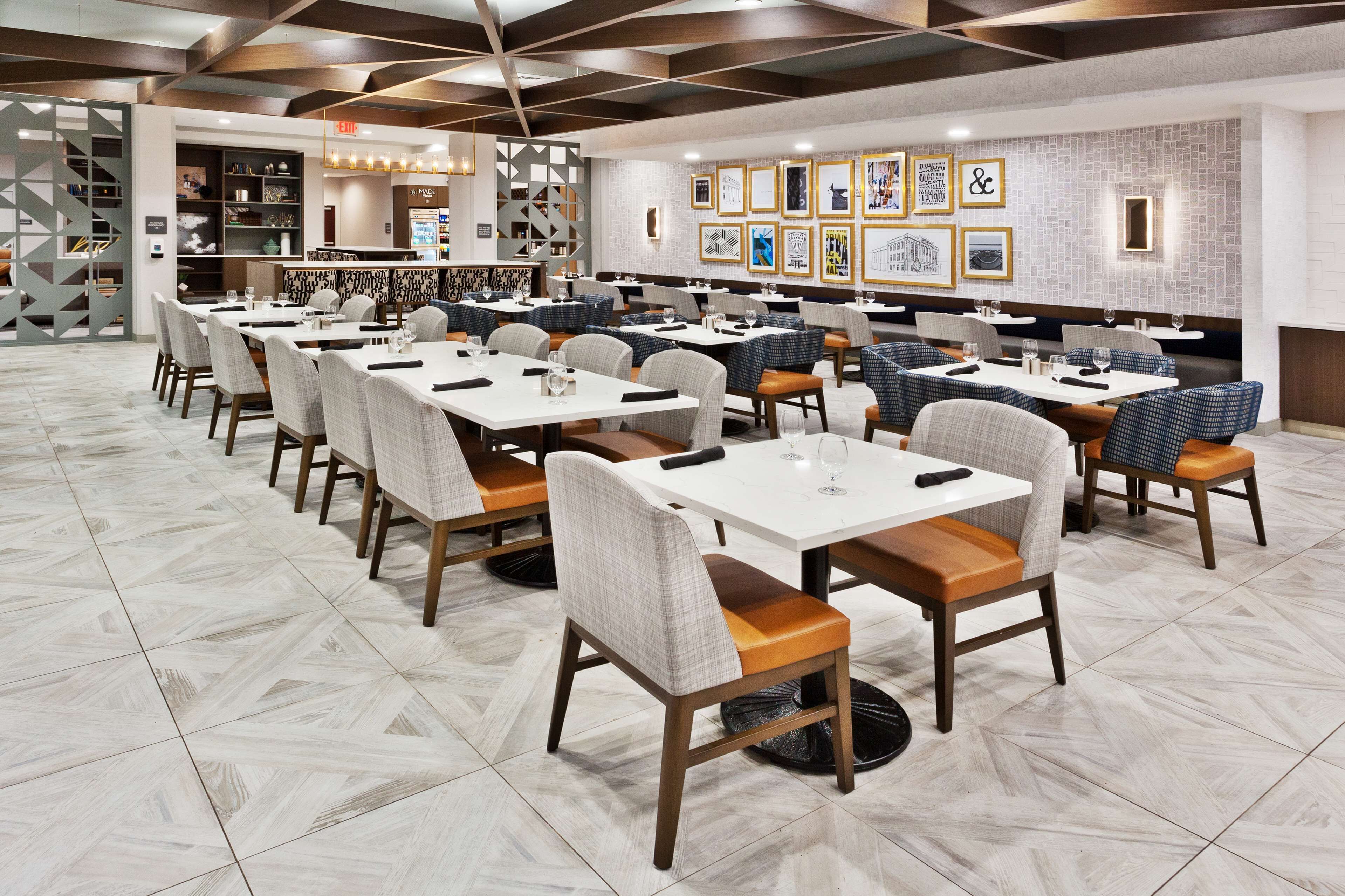 A modern restaurant with multiple dining tables and chairs, a bar area, and a wall decorated with framed pictures.