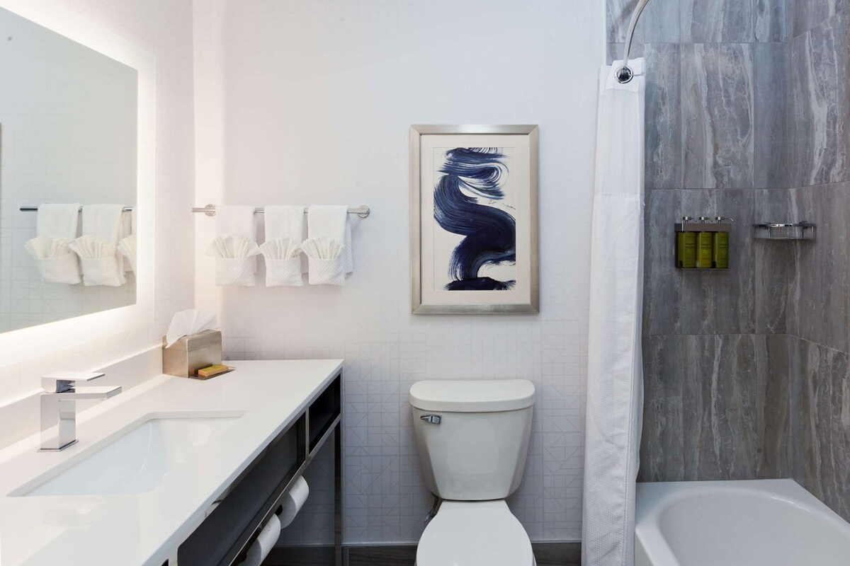 Une salle de bain moderne avec un meuble-lavabo blanc, des toilettes, une douche aux carreaux gris et une œuvre d’art encadrée au mur.