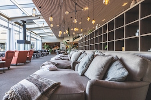 Un área de descanso moderna con sofás y sillones, techo de madera con luces colgantes, y un ventanal que proporciona luz natural.