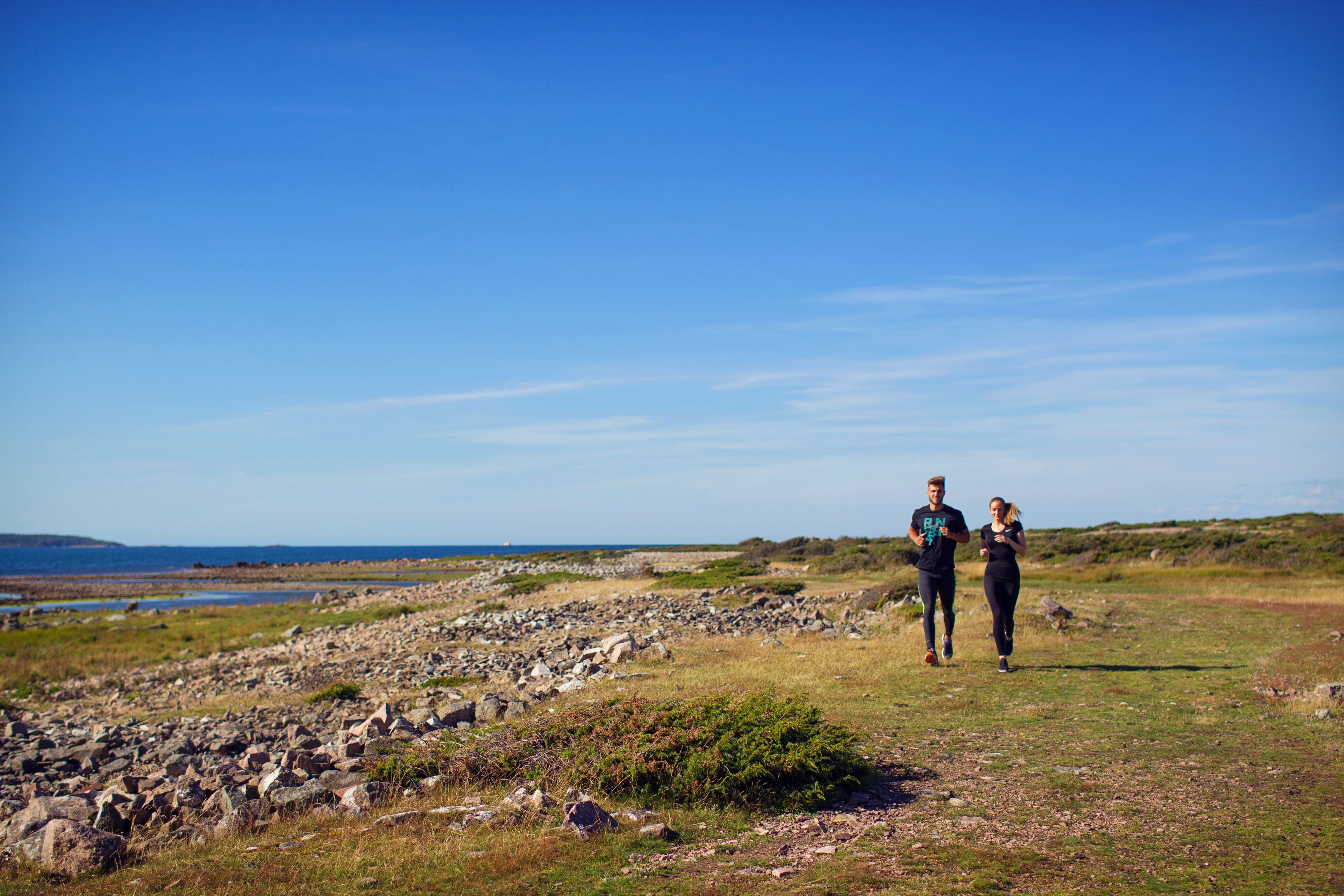 Två personer som joggar på en stenig stig med utsikt över havet och kullarna i bakgrunden.