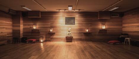 A room with wooden panel walls, a central meditation area with a statue, and four wall-mounted lamps.
