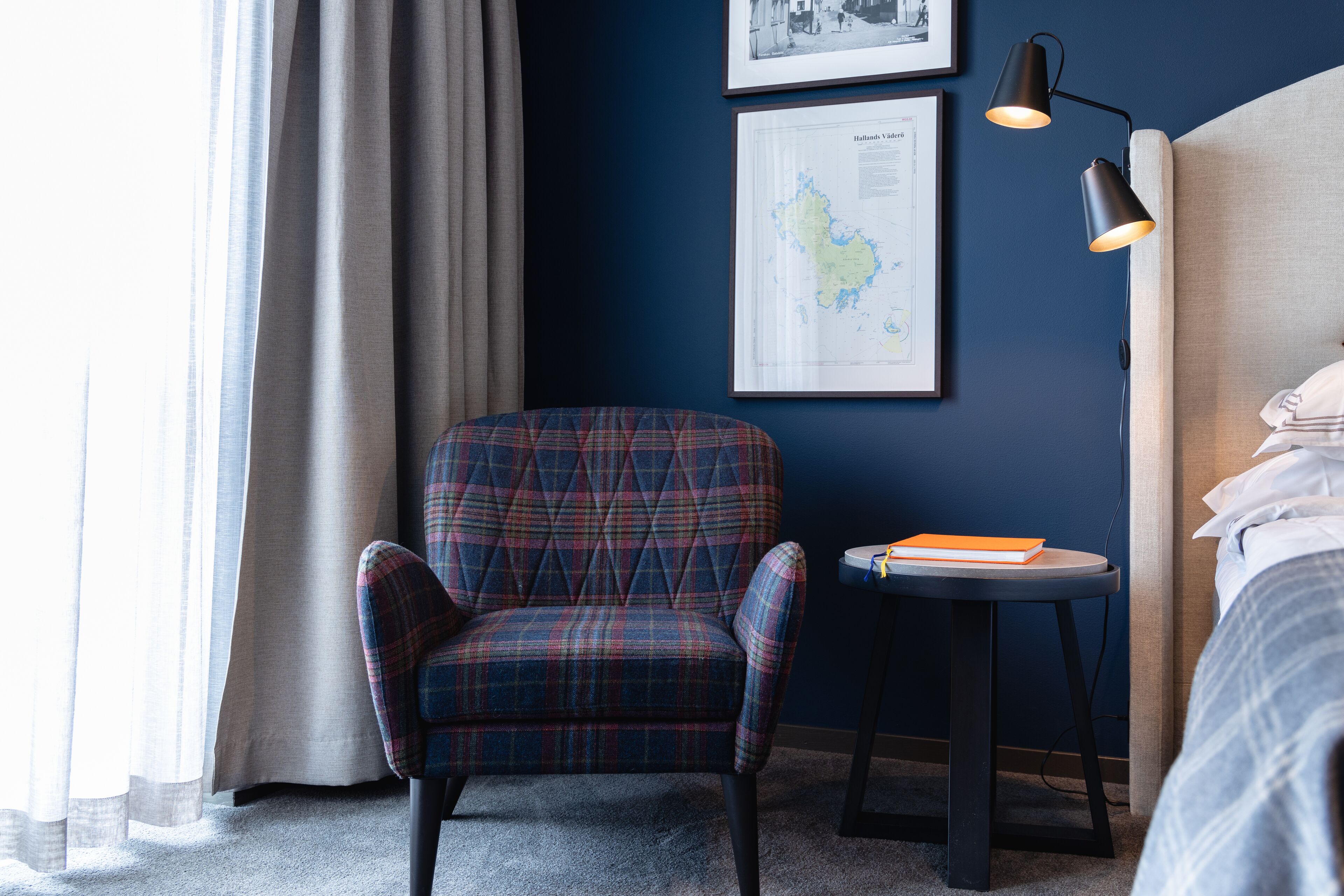 A hotel room with a plaid armchair, a small table with a book, a map on the wall, and a bed with white linens.