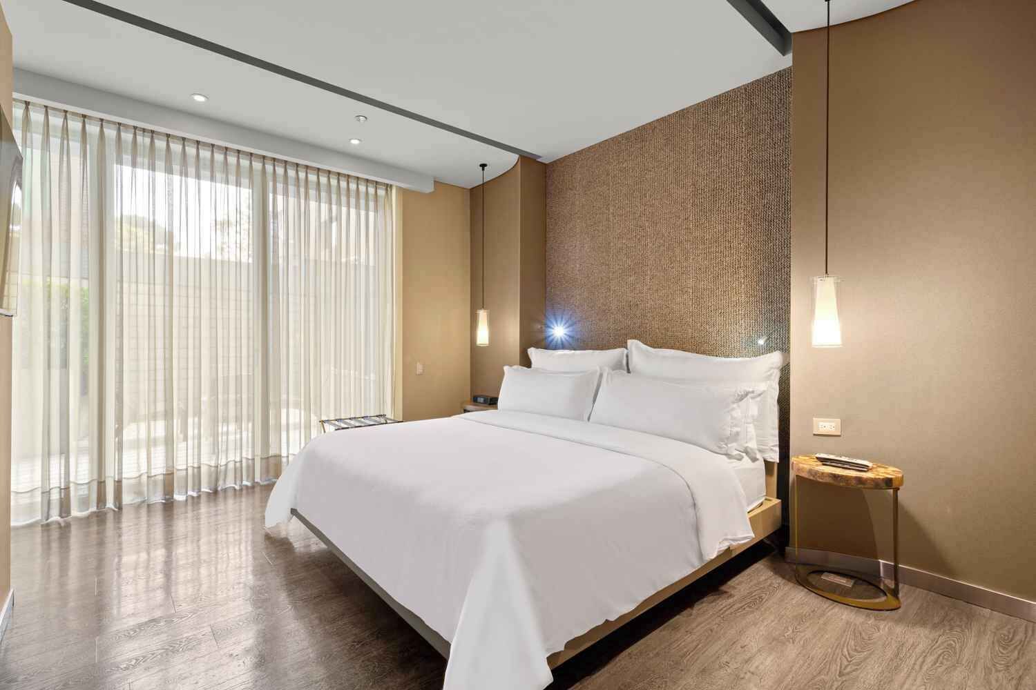 Habitación de hotel moderna con una cama grande, una mesita de noche, una lámpara y vistas a través de cortinas translúcidas.