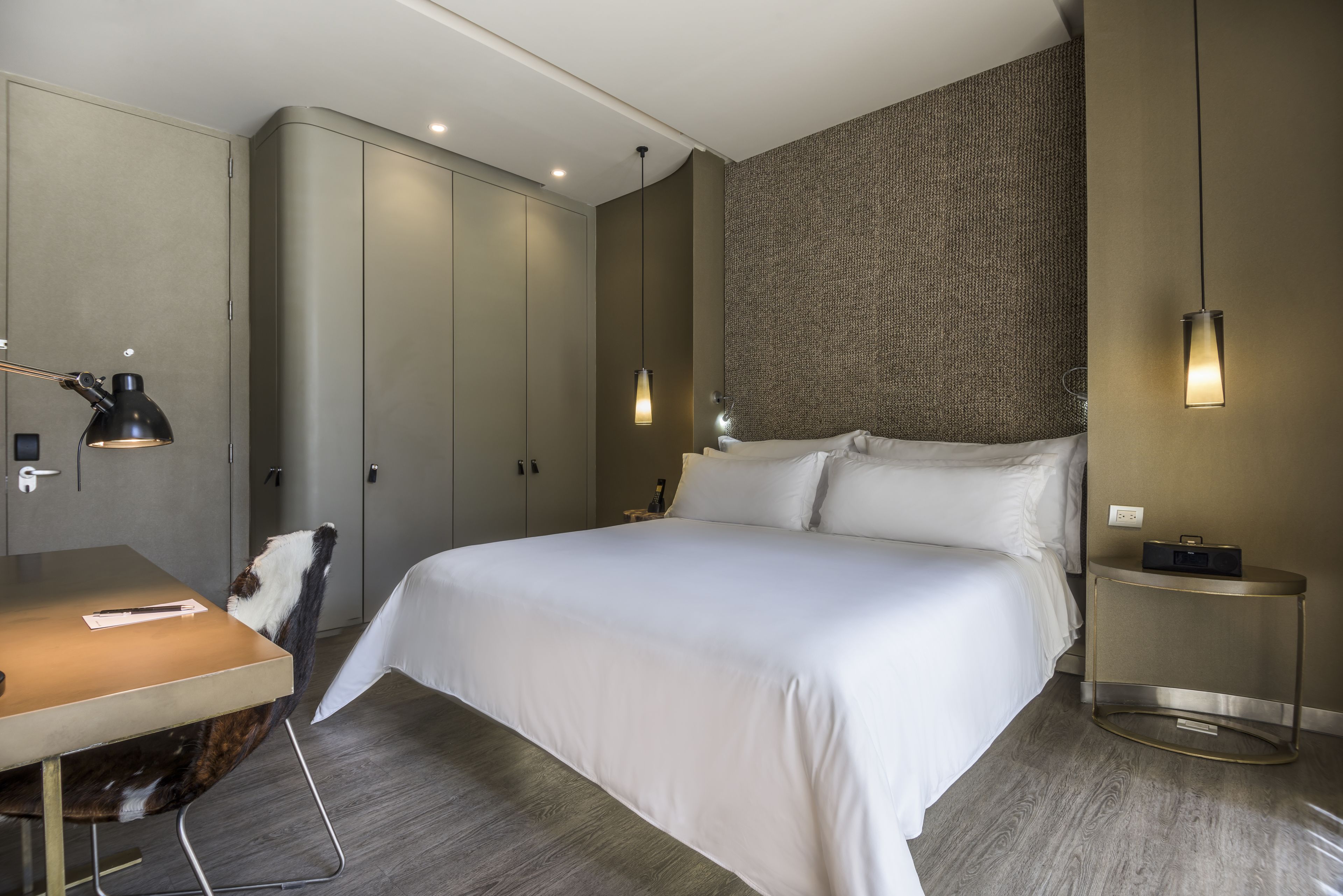 A modern hotel room with a large bed, a desk with a chair, a wardrobe, and two bedside tables with lamps.