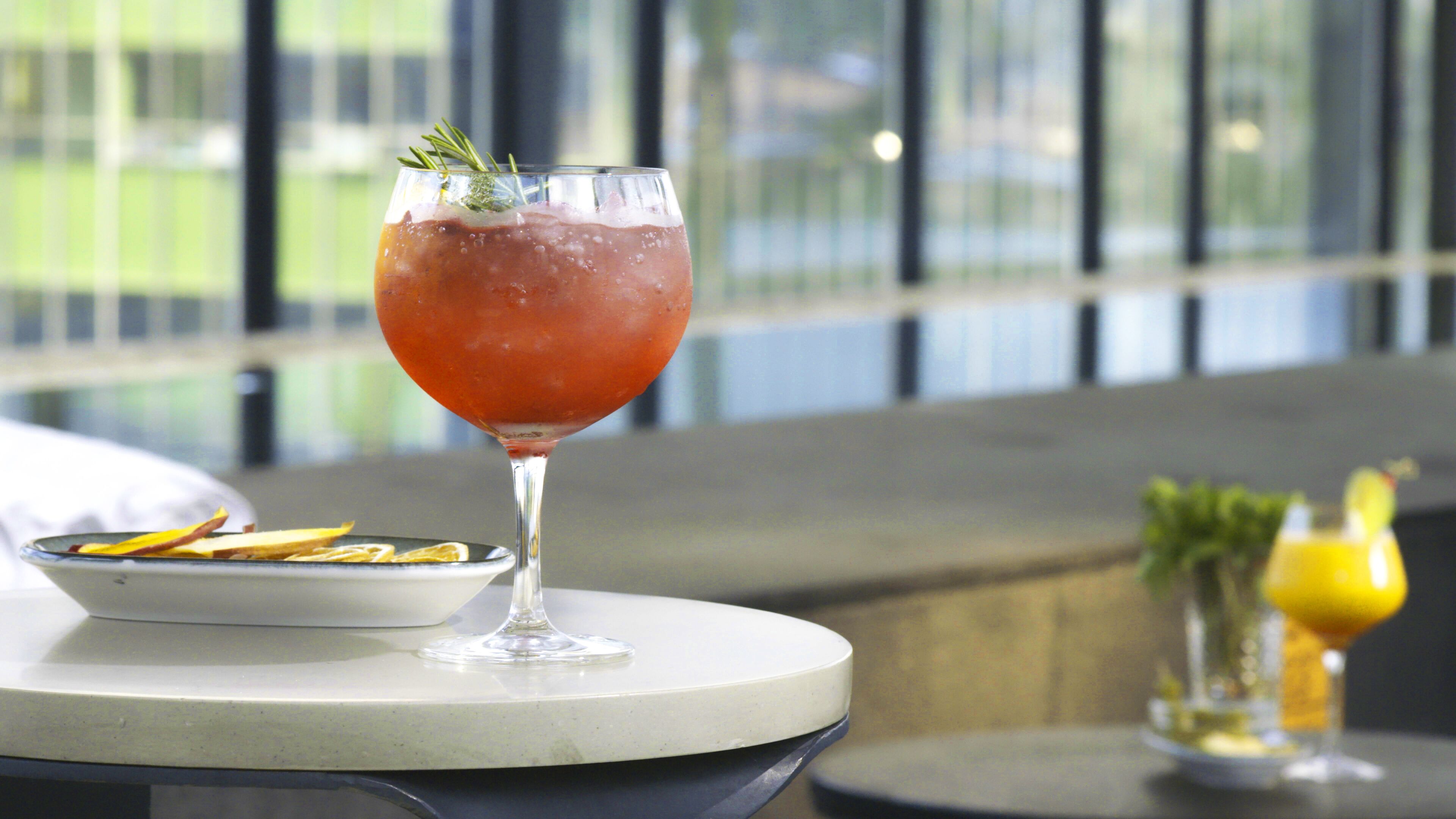 A glass of red drink with a garnish on a table with a plate of food and a small bowl.