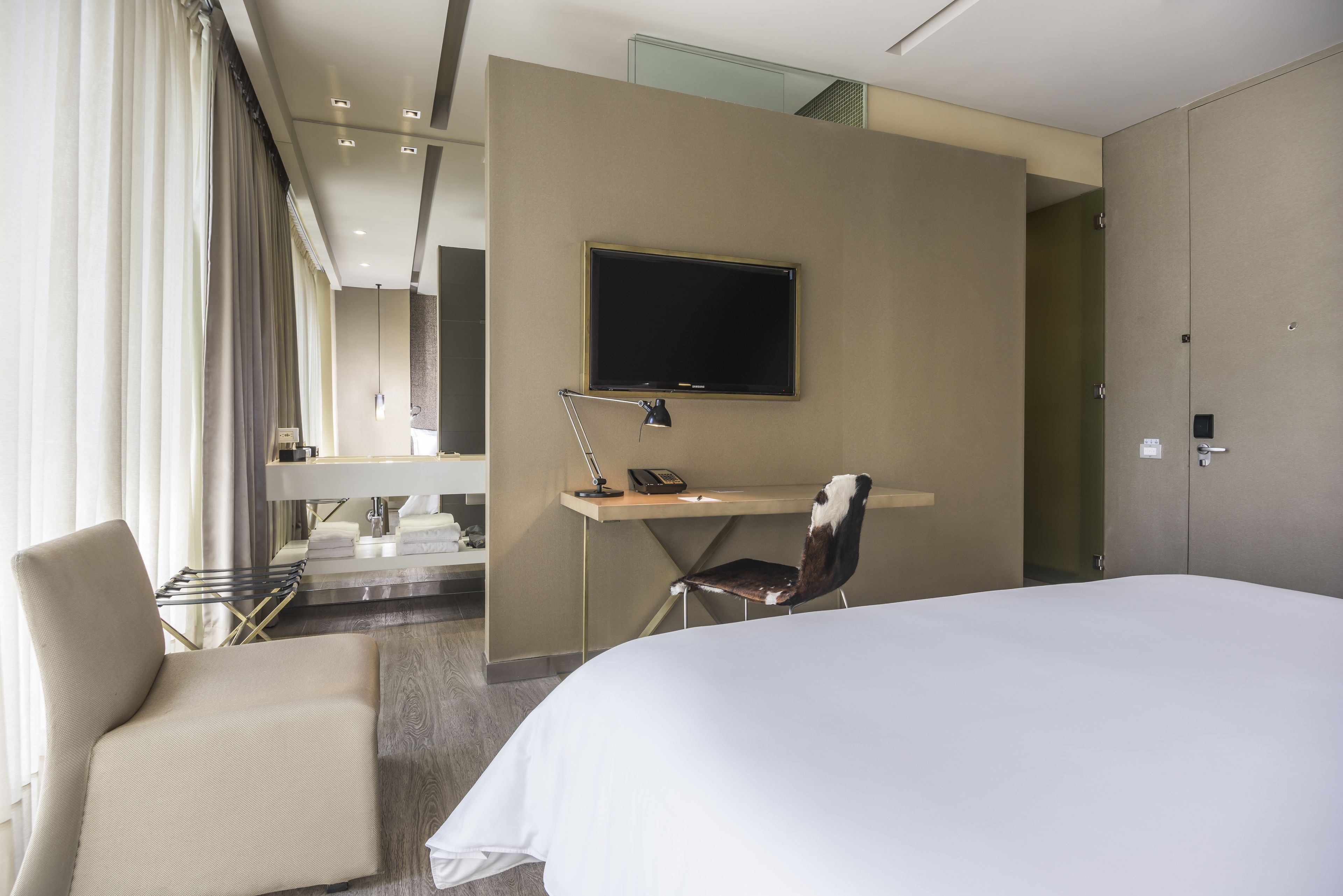 Habitación de hotel con una cama, un escritorio con silla, un televisor montado en la pared y una ventana con cortinas.