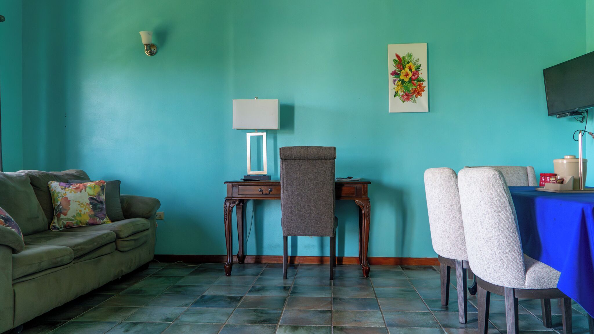 A room with teal walls, a sofa, a desk with a chair, a TV, and a table with chairs.