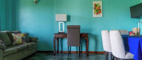 A room with teal walls, a sofa, a desk with a chair, a TV, and a table with chairs.