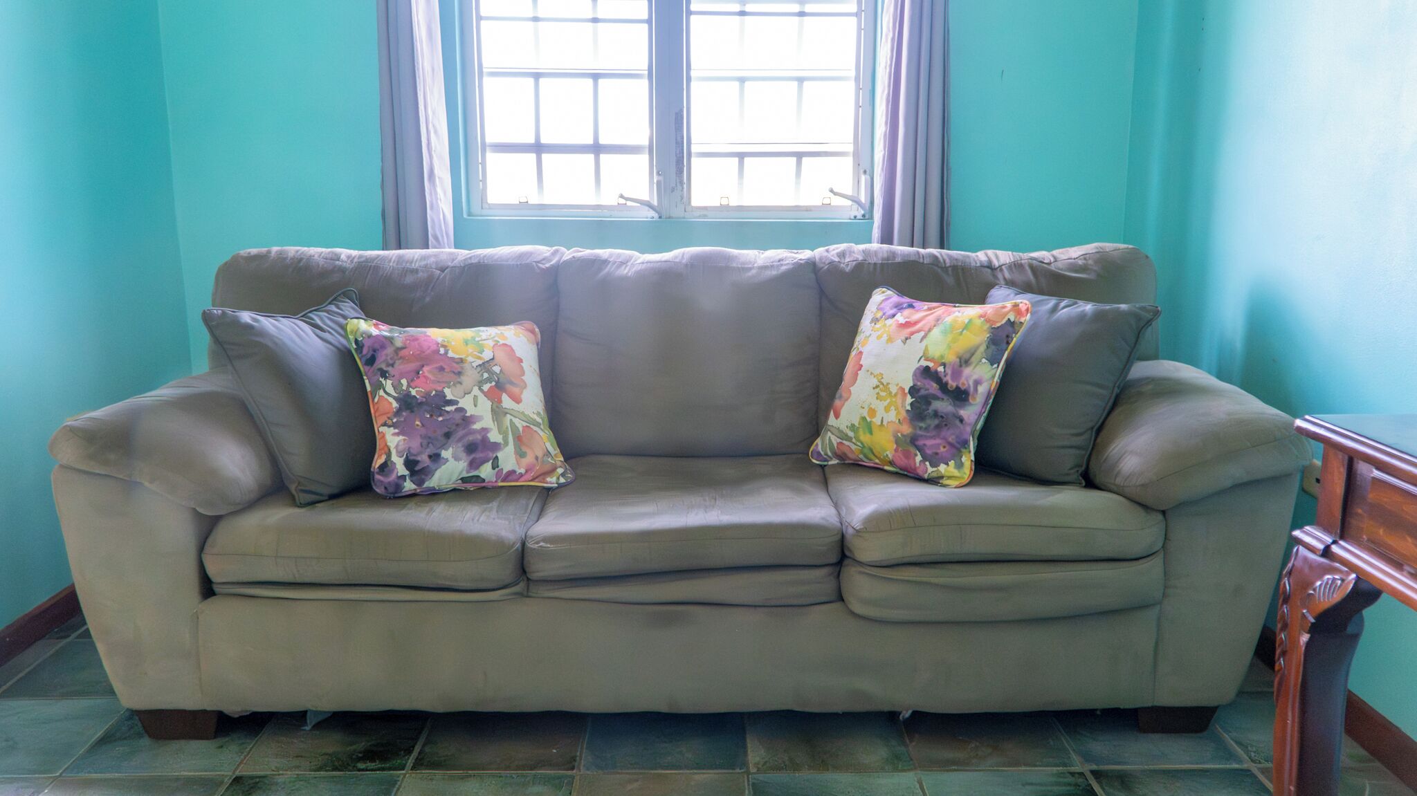 A beige leather sofa with two colorful floral cushions, positioned in front of a window with a view of a building.