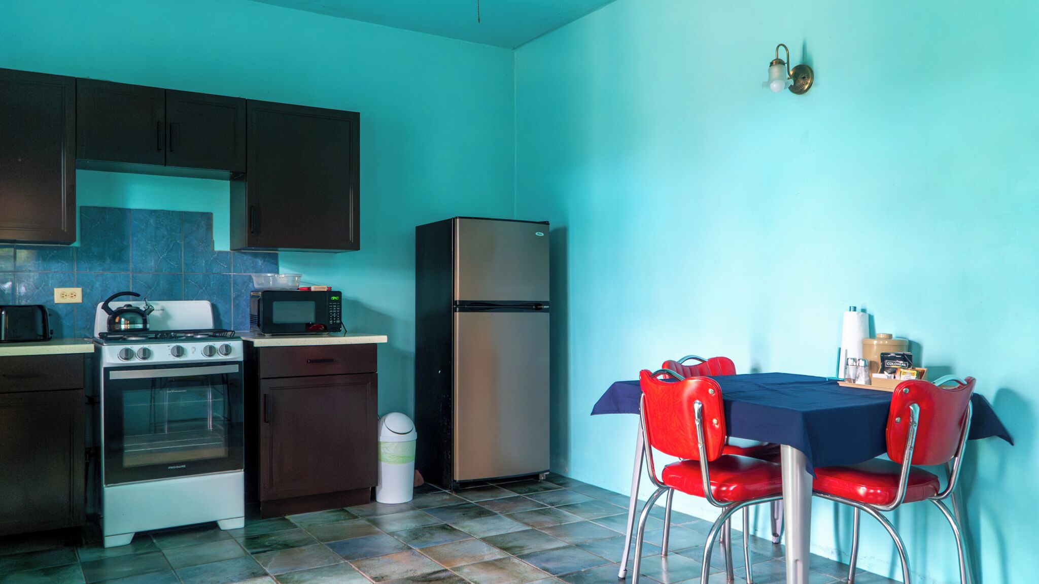 Una cucina compatta con fornello, forno, microonde e frigorifero. Si nota un tavolo da pranzo con sedie rosse e una tovaglia blu. Vicino alla cucina, è presente un cestino.