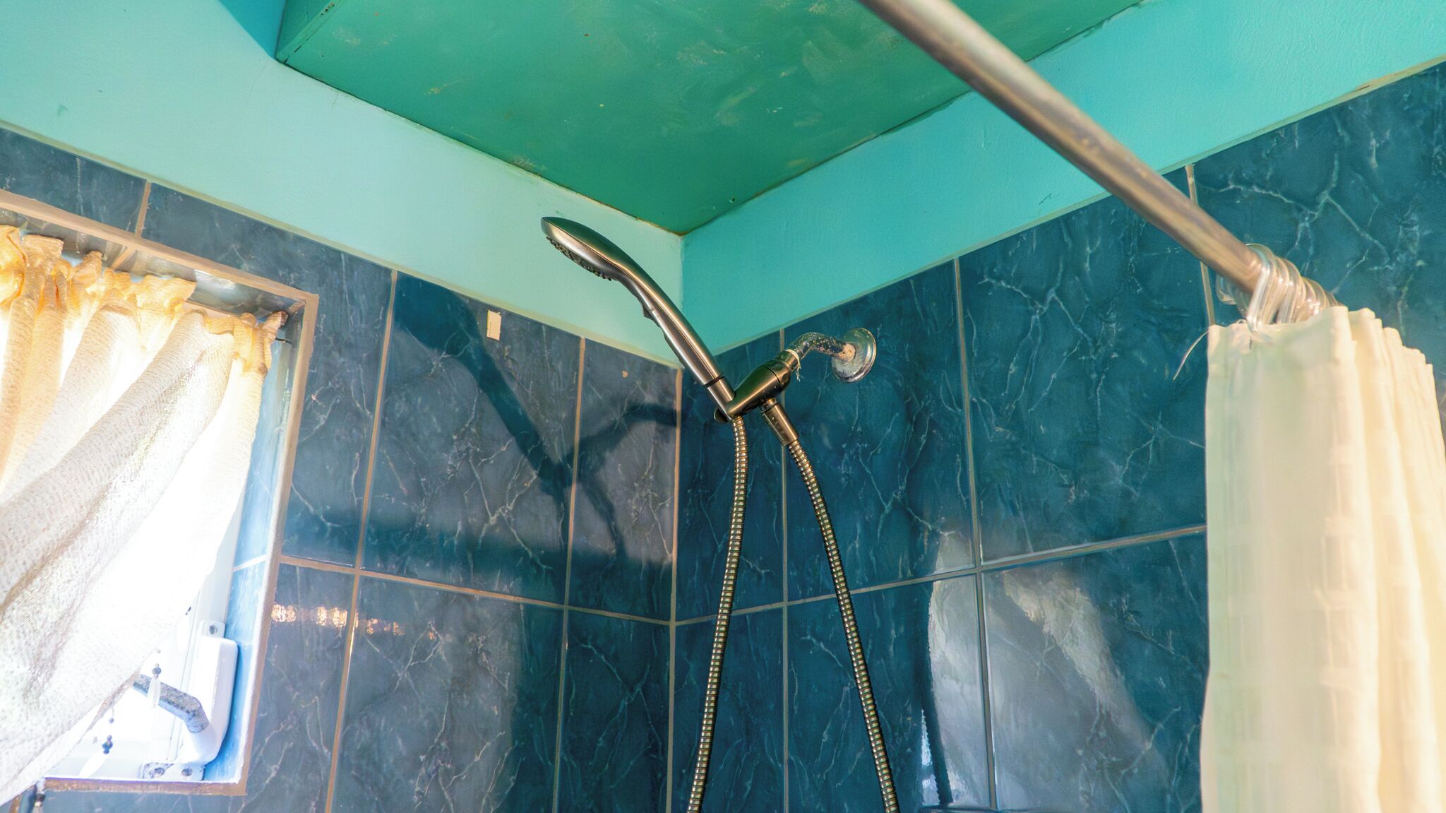 A bathroom with dark blue tiles, a shower with a handheld showerhead, and a window with a curtain.