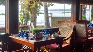 A dining area with a table set for two, wooden chairs with woven backs, and a view of a body of water through large windows. - Coconut Lagoon Kumarakom- A CGH Earth Experience (Kottayam)