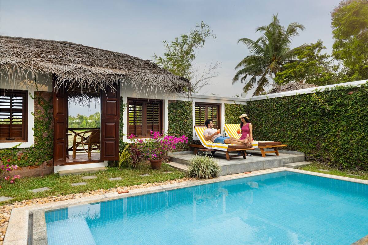 Un espace aménagé au bord de la piscine, comprenant une structure à toit de chaume, une piscine et un jardin agrémenté de plantes en pot.