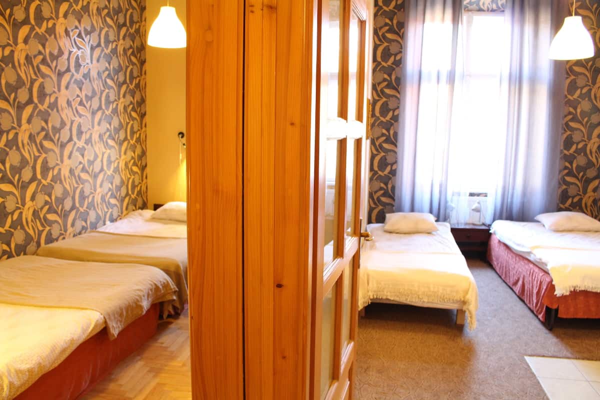 A hotel room with three single beds, wooden flooring, patterned wallpaper, and a large window with curtains.