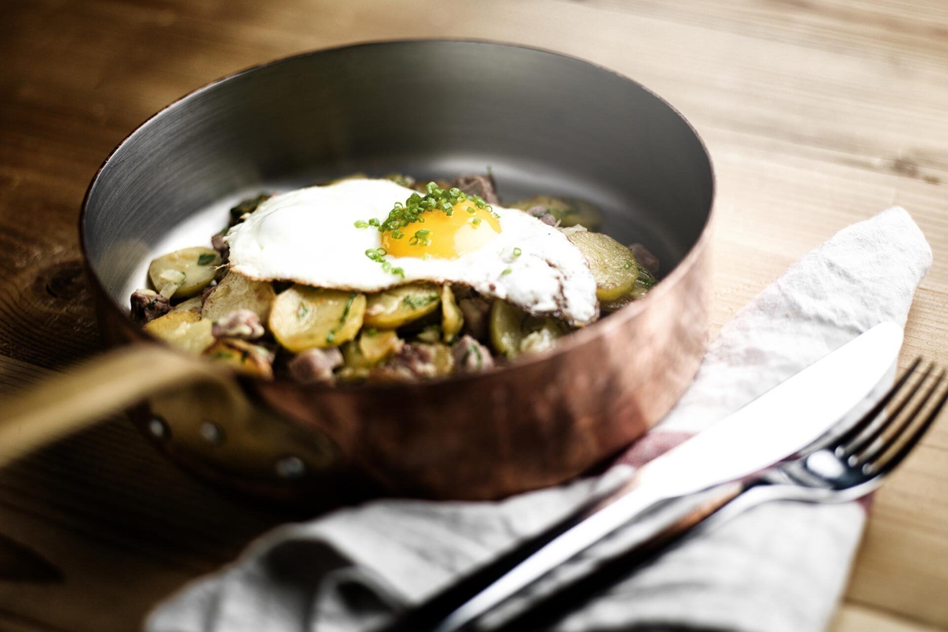 A skillet with a fried egg and potatoes, a knife, and a fork on a wooden table.