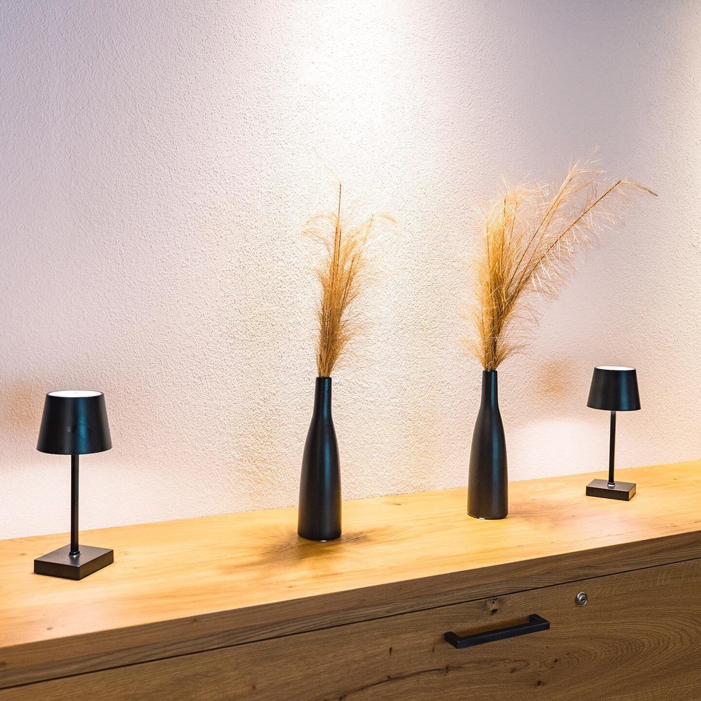 Eine Holzkommode mit drei schwarzen Lampen und zwei Vasen mit getrockneten Pflanzen.