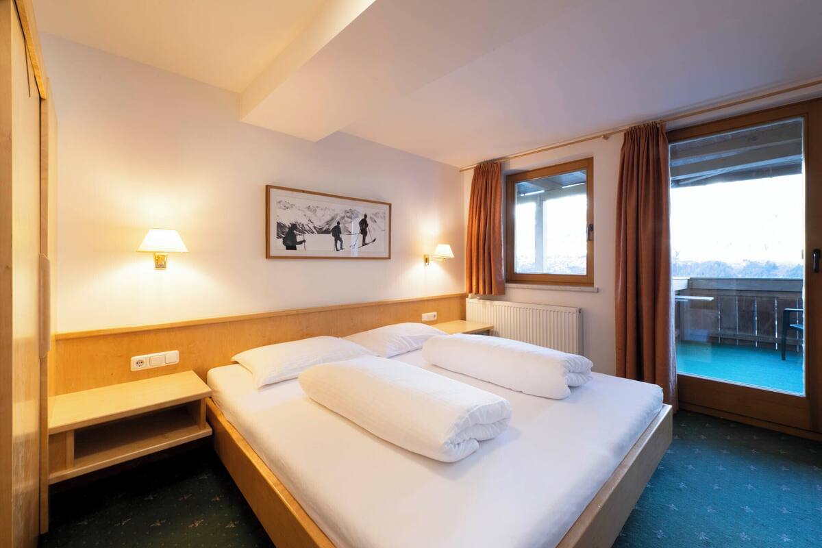 Habitación de hotel con cama, dos mesitas de noche, ventana con cortinas, radiador y un cuadro en la pared.