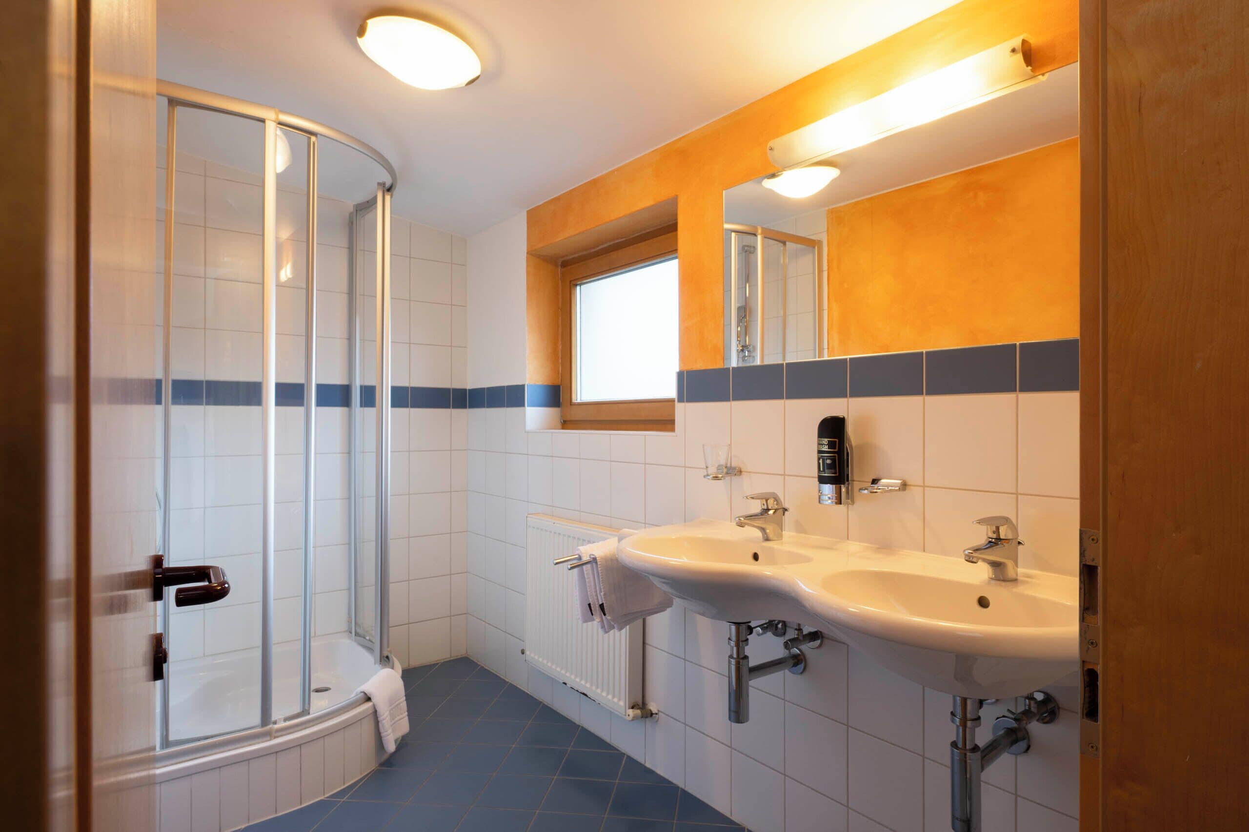 A bathroom with a glass shower enclosure, two sinks, a mirror, and a window.