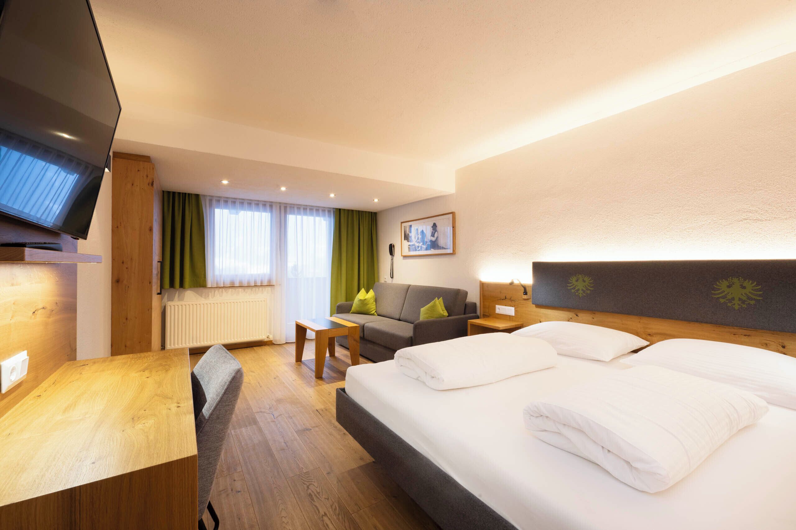 A hotel room with a bed, a desk, a chair, a sofa, a TV, and a window with curtains.