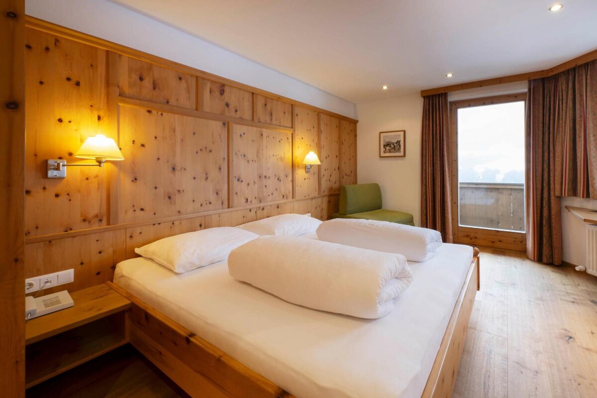Habitación de hotel con cabecera de madera, dos lámparas de noche, una silla verde, cama con ropa blanca y una ventana con cortinas.
