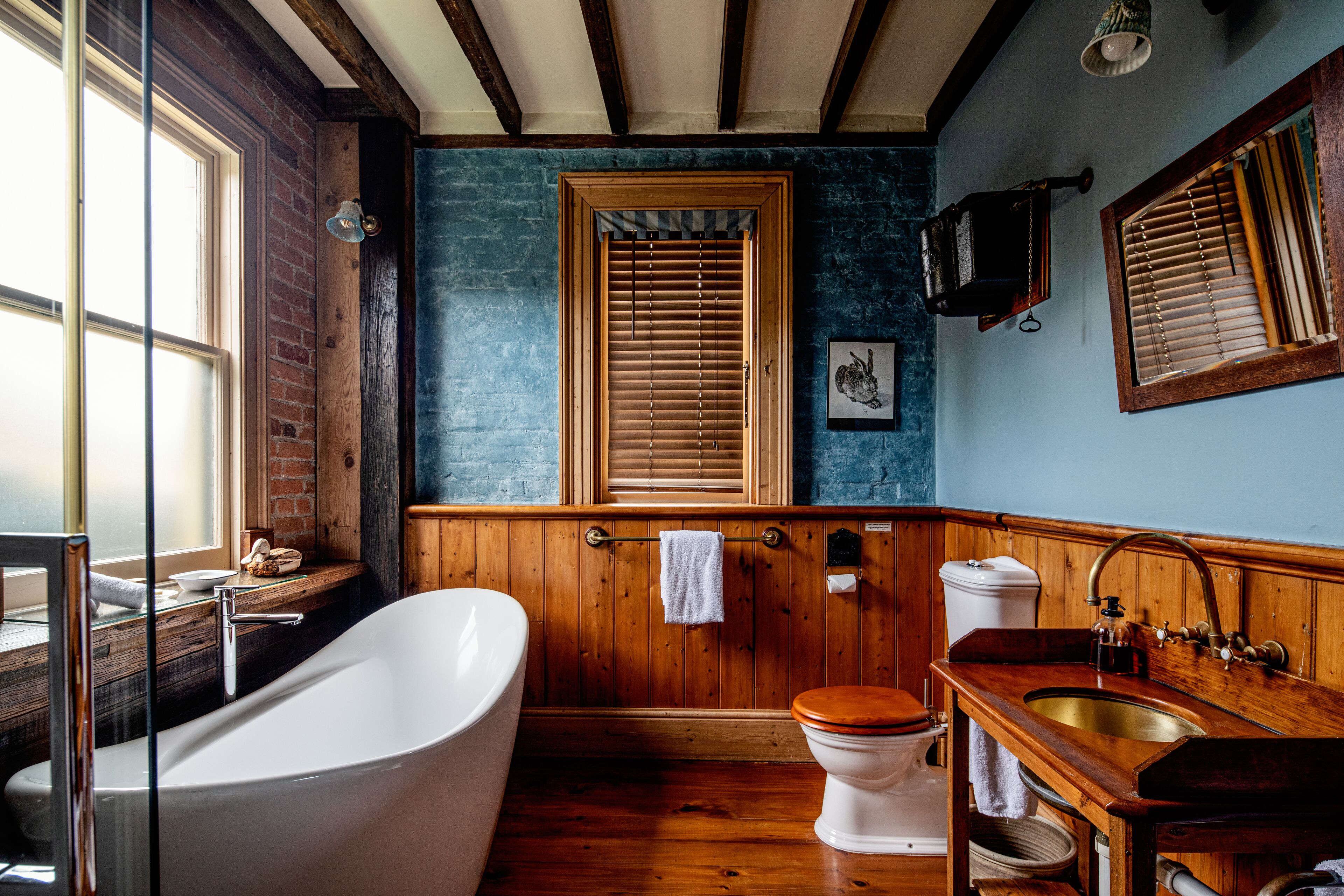 A bathroom with a freestanding bathtub, a wooden vanity, a toilet, and a window with blinds.