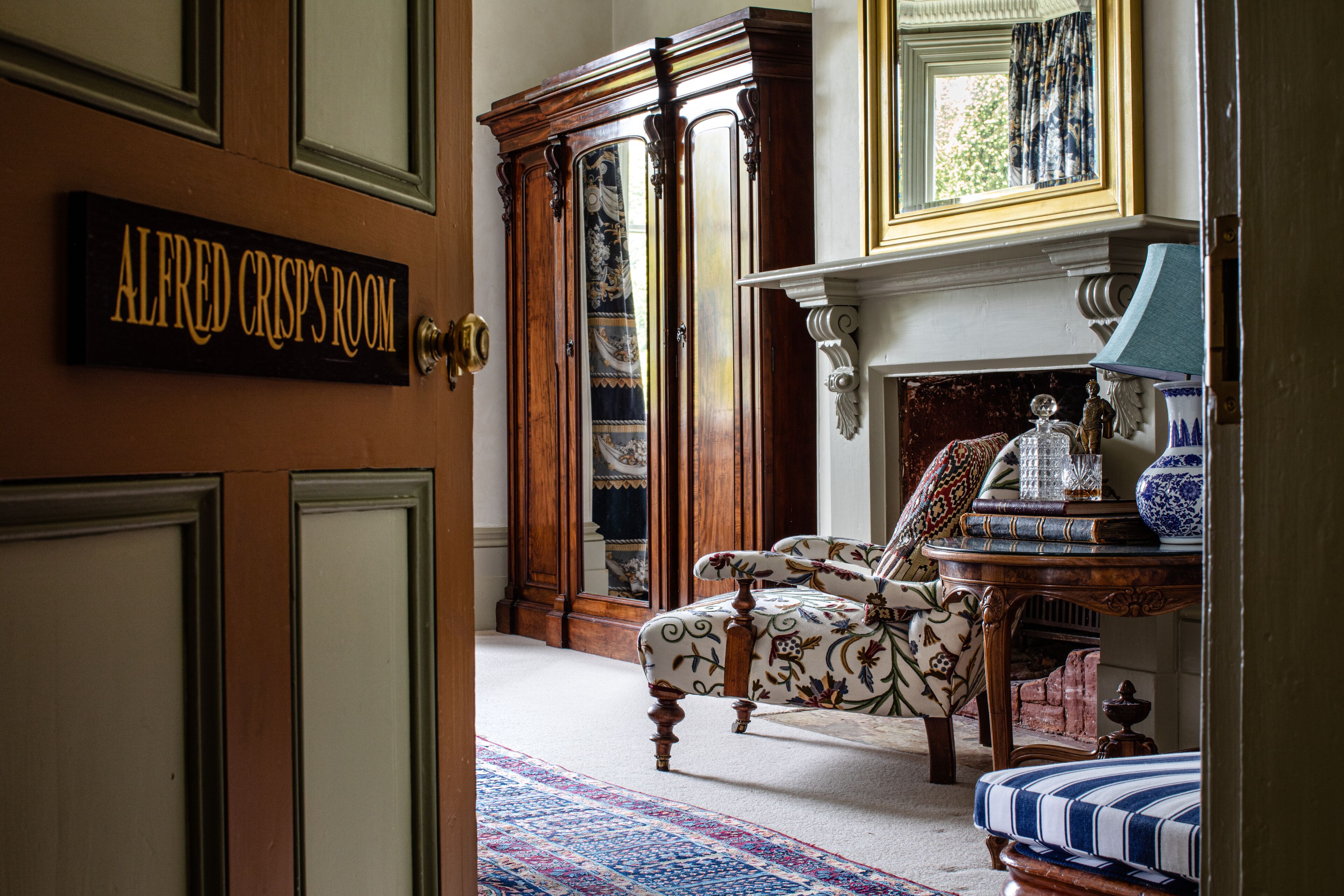 Una habitación con una puerta de madera que dice "ALFRED CRISP'S ROOM", un sillón estampado, un armario de madera y una mesa con una lámpara y objetos decorativos.