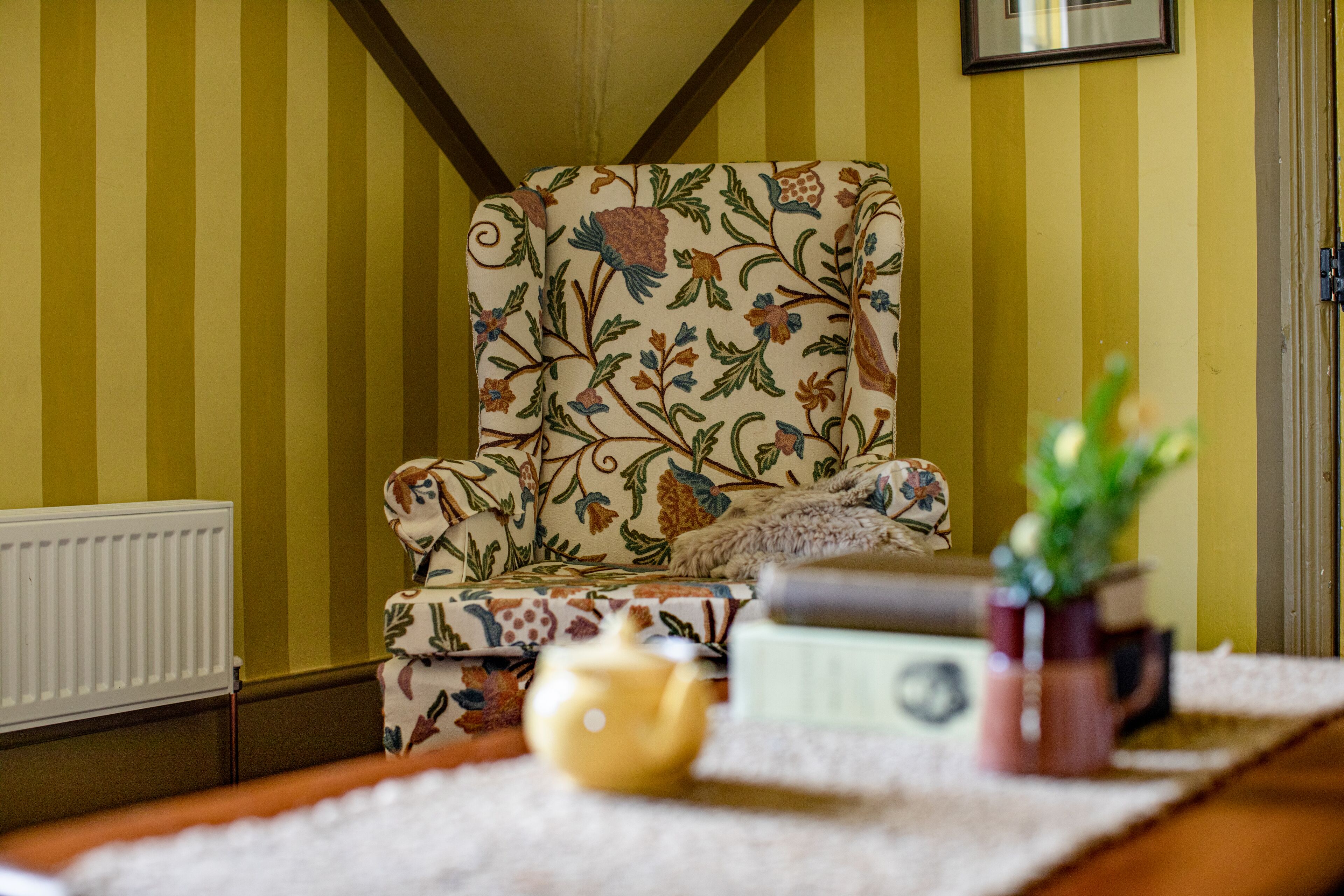 Une pièce avec un mur à rayures, un fauteuil à motifs floraux, un radiateur, une table avec une plante, une théière et une tasse.
