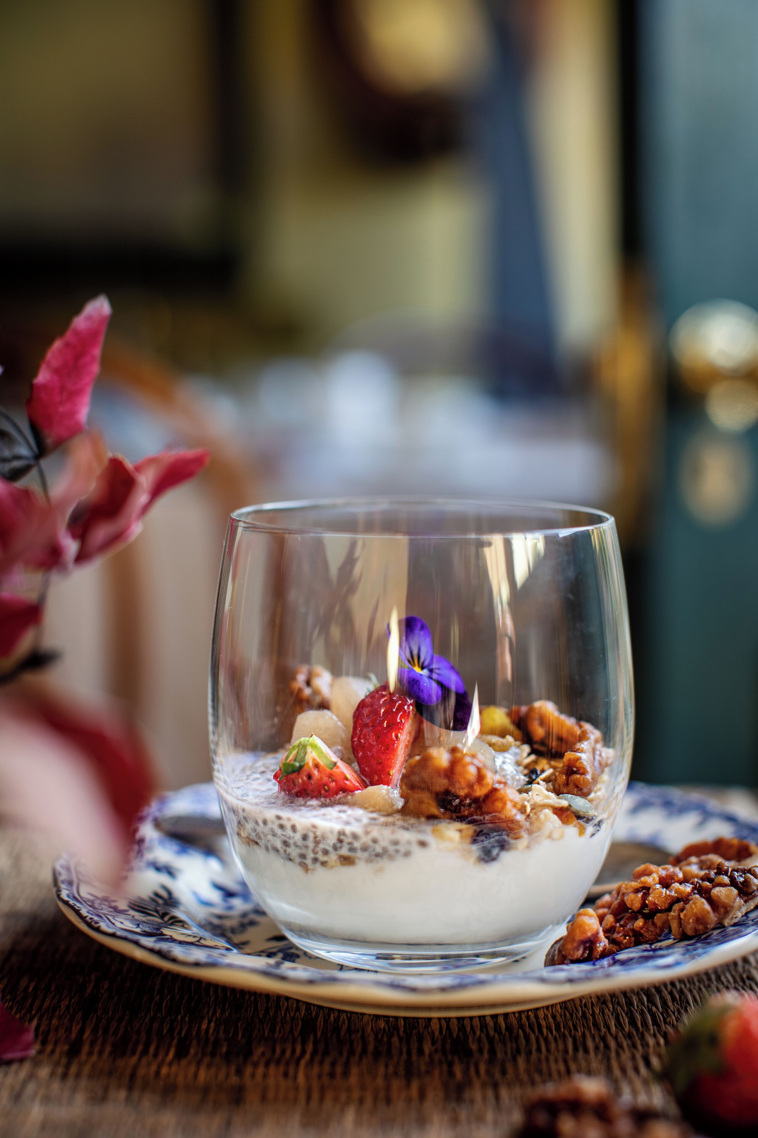 A clear glass bowl with a layered dessert, including a strawberry, a purple flower, and granola, on a patterned plate.