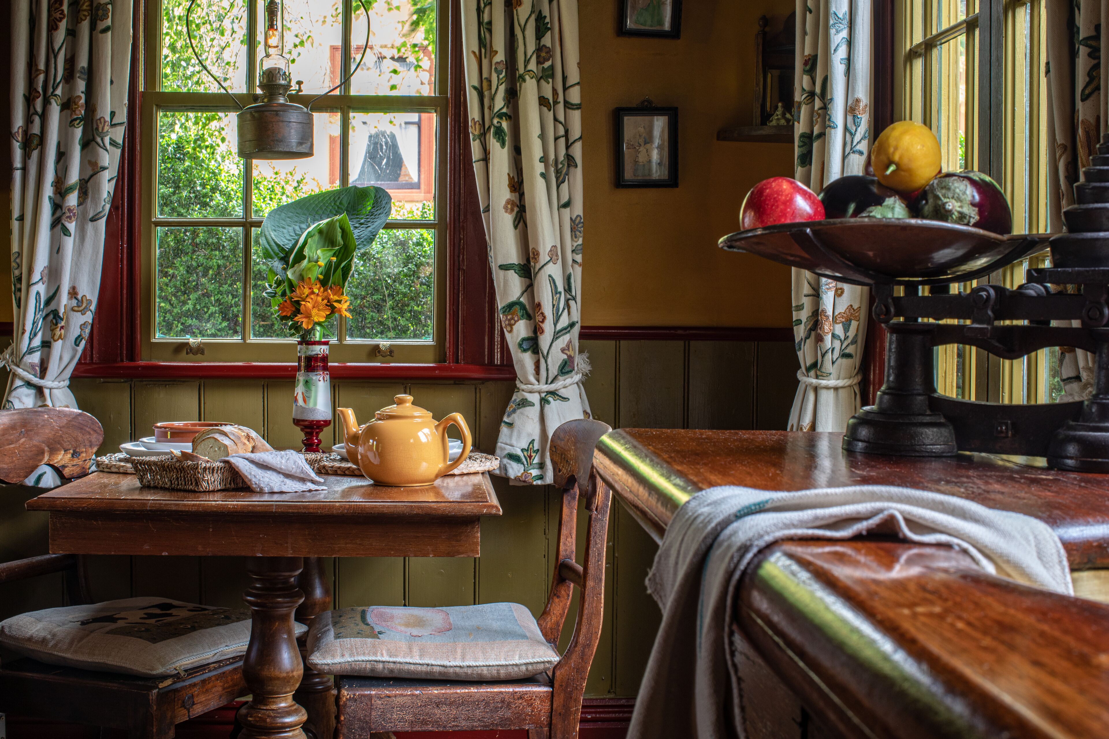 A cozy room with a wooden table, a window with curtains, and a fruit bowl on a stand.