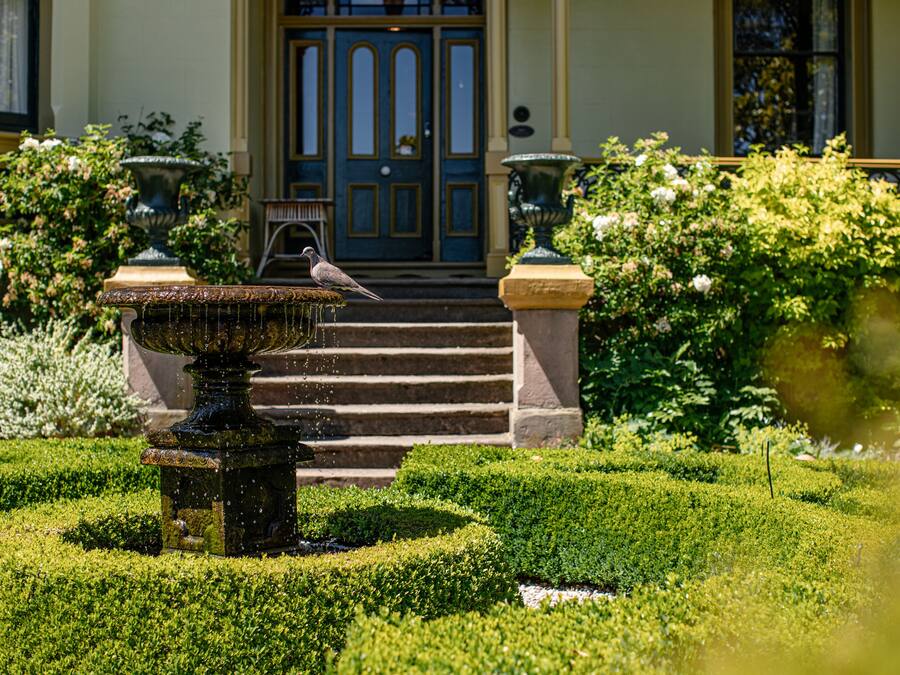A well-manicured garden with a fountain, steps leading to a door, and a building with a blue door in the background.
