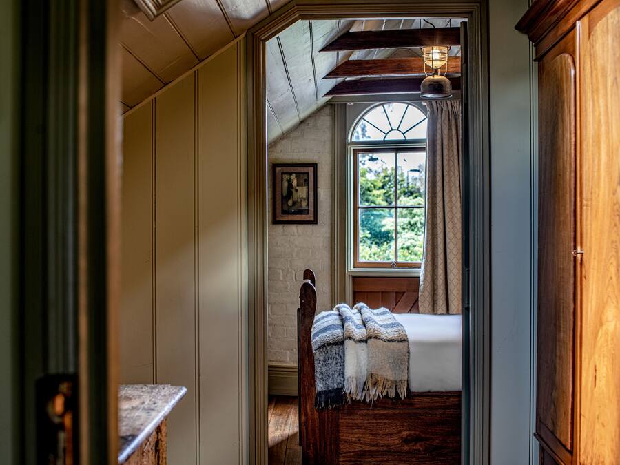 A narrow hallway leading to a bedroom with a wooden bed, a window with curtains, and a framed picture on the wall.