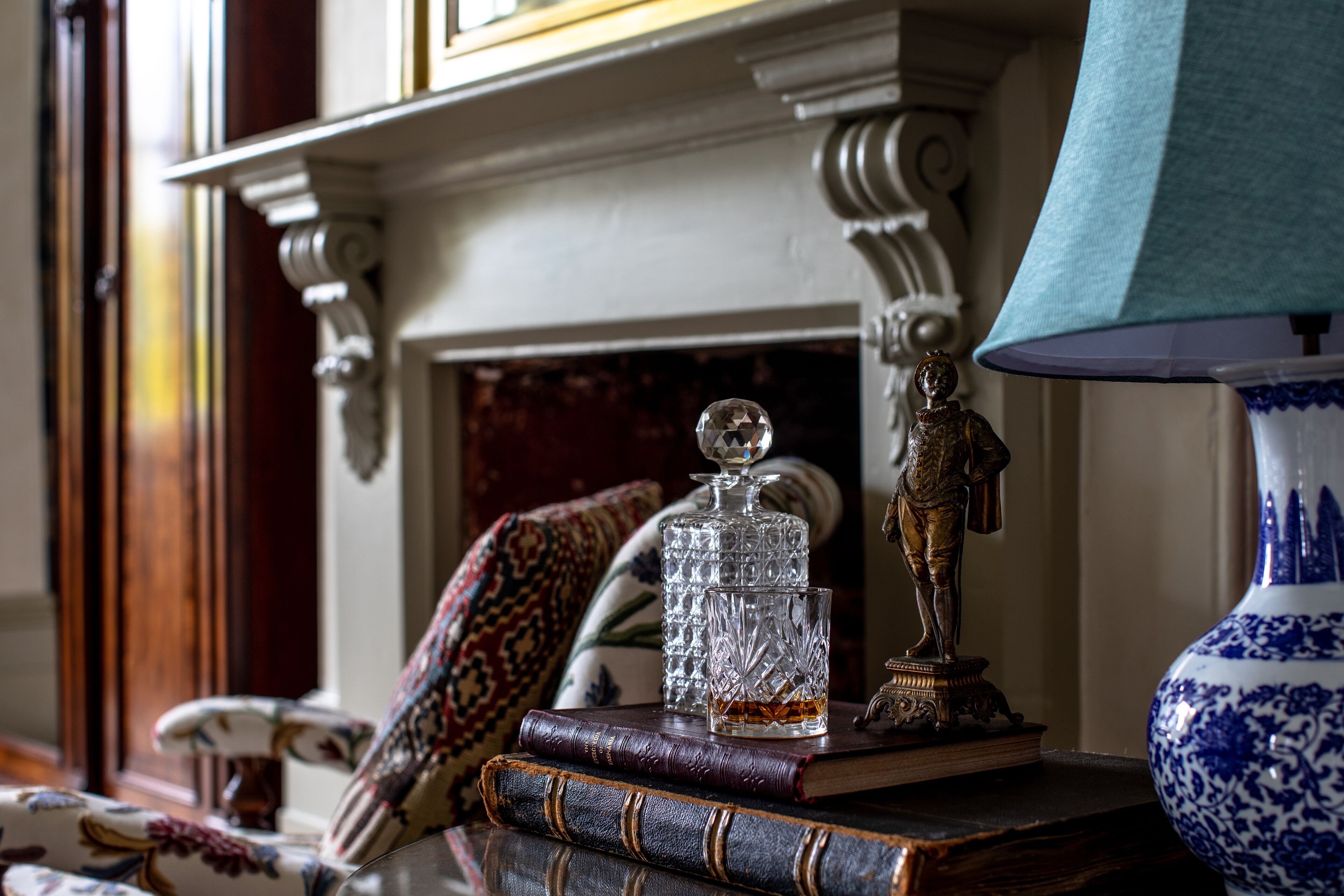 A fireplace with ornate mantel, a crystal decanter, a brass statue, and a blue and white vase on a table.